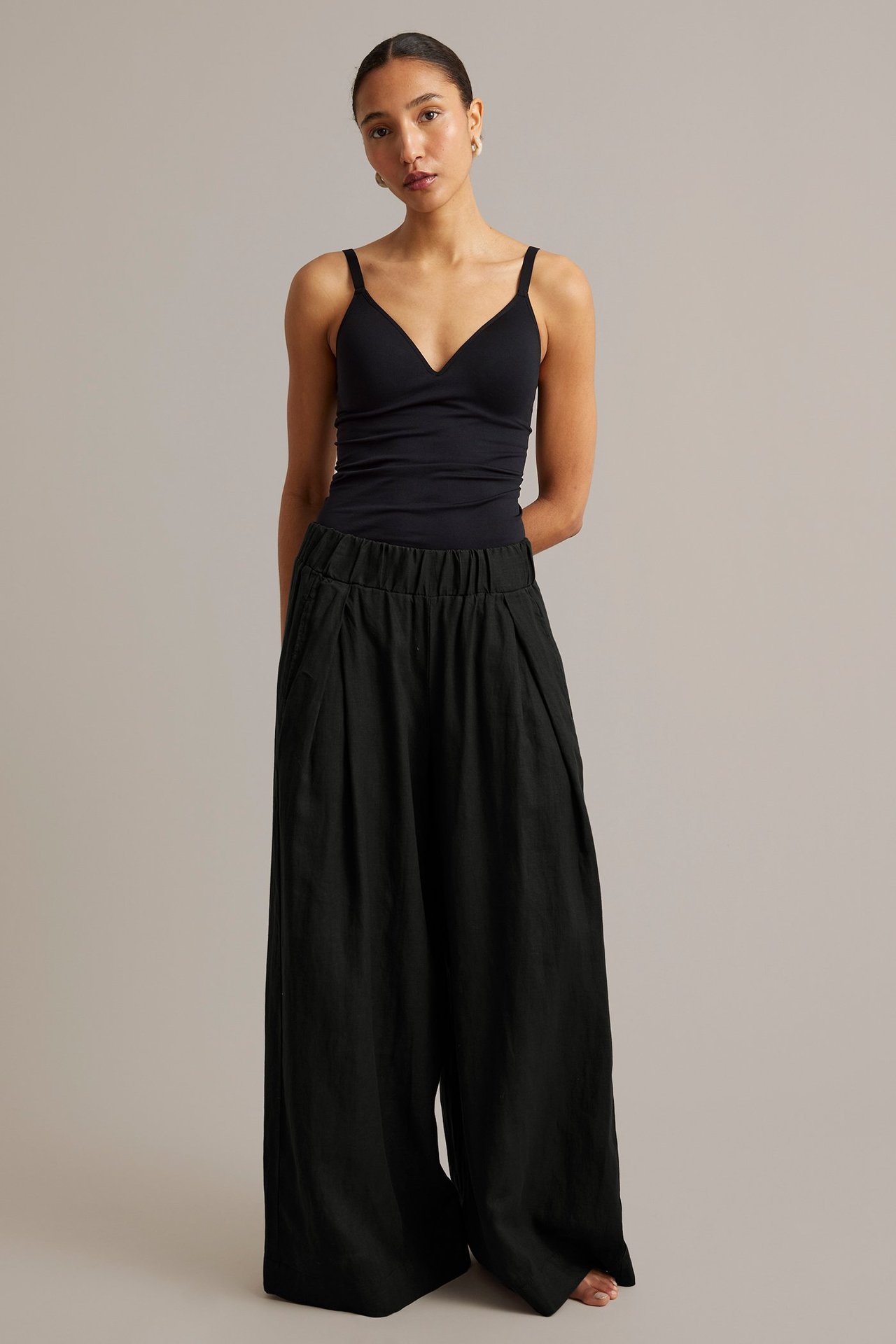 Nainen mustissa leveälahkeisissa pellavahousuissa joustavalla vyötäröllä, laskoksilla ja mustassa V-aukkoisessa topissa.