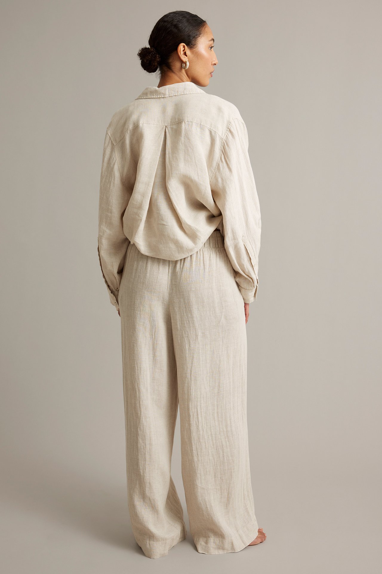 Bakfra: kvinne i løstsittende beigemelert vid linbukse og matchende langermet skjorte.