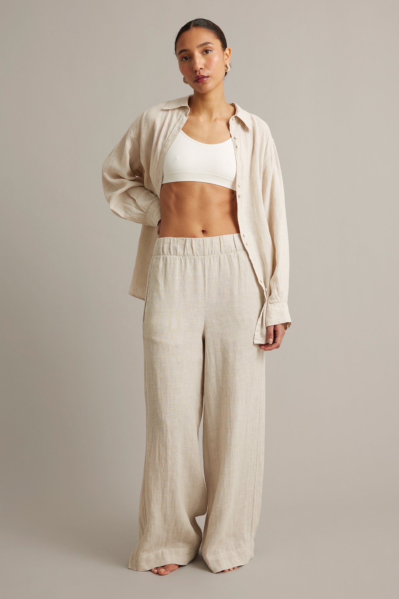 Frontbilde av kvinne i beige melert linbukse med vide ben, hvit BH og åpen beige linskjorte.