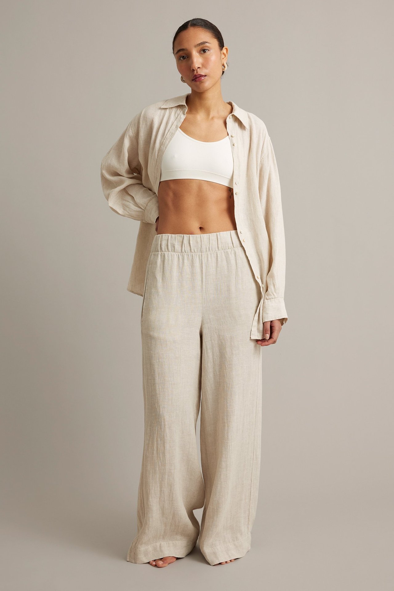 Naisen etukuva beigeissä meleratuissa pellavahousuissa, valkoisissa rintaliiveissä ja avoimessa pellavapaidassa.