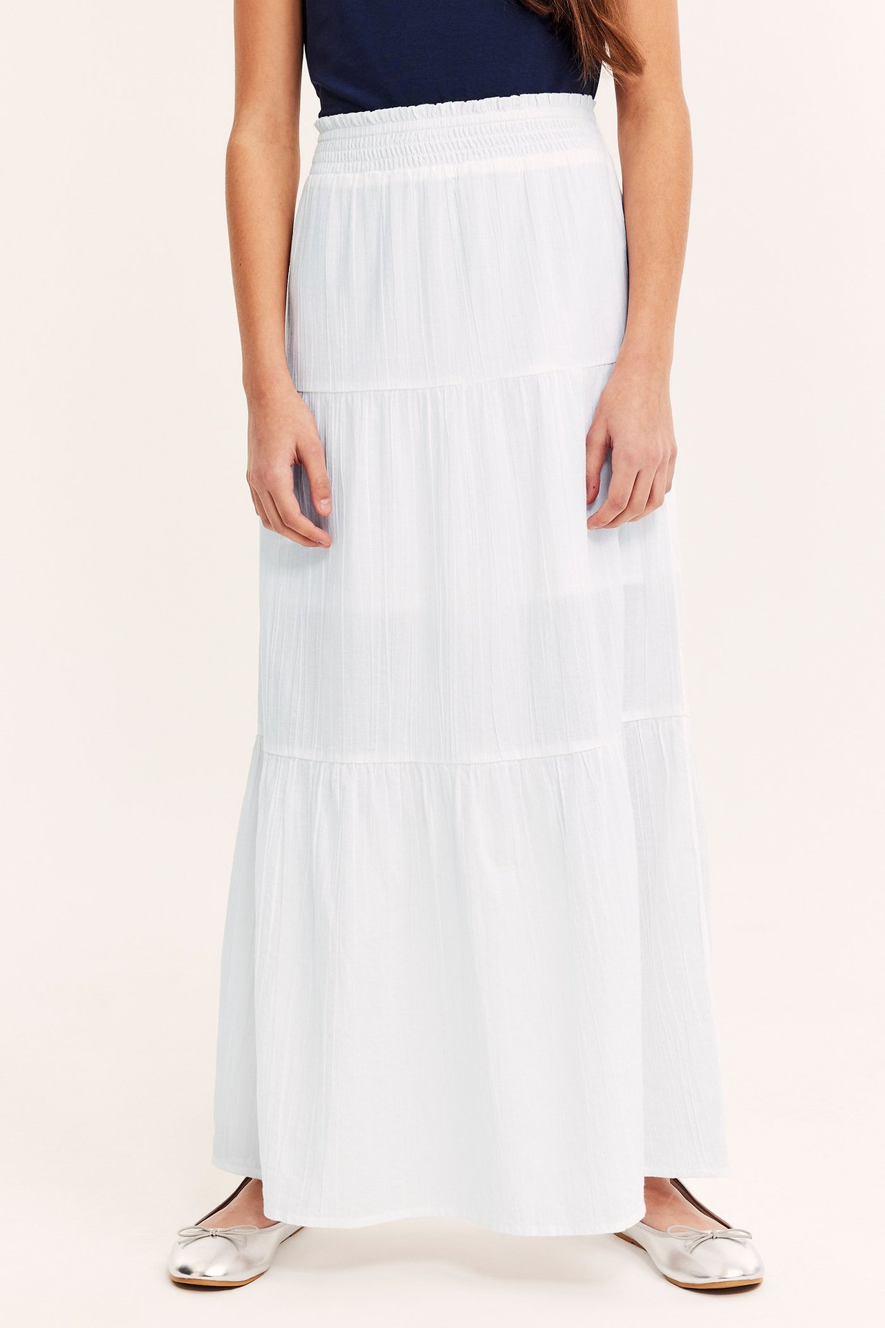 Frontvisning av et barn i et hvitt lagdelt maxiskjørt, mørkeblå t-skjorte og sølvfargede ballerinasko.