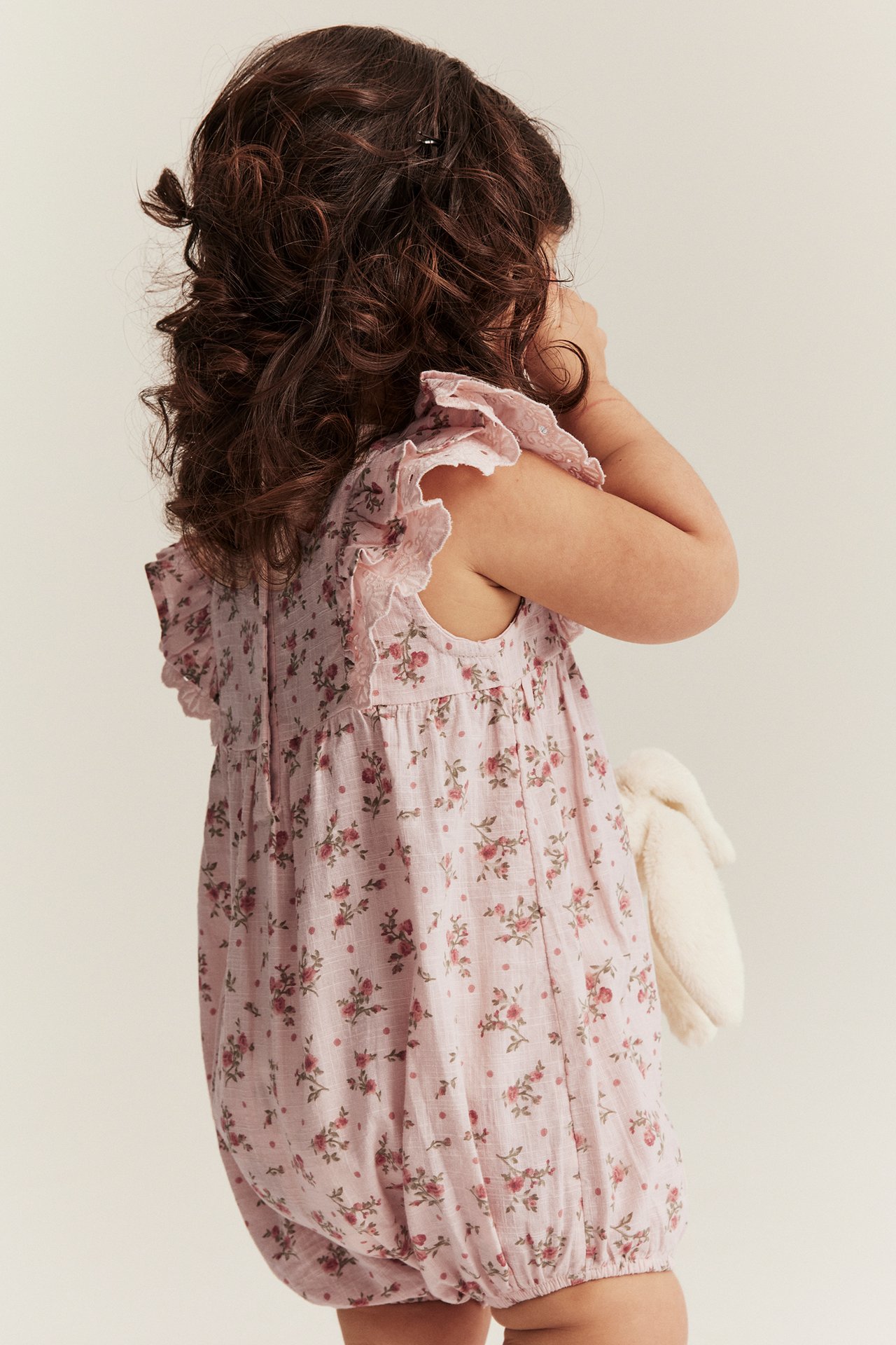 Sivukuva vauvasta vaaleanpunaisessa kukkahaalarissa, jossa röyhelöhihat, pitäen valkoista pehmolelua.