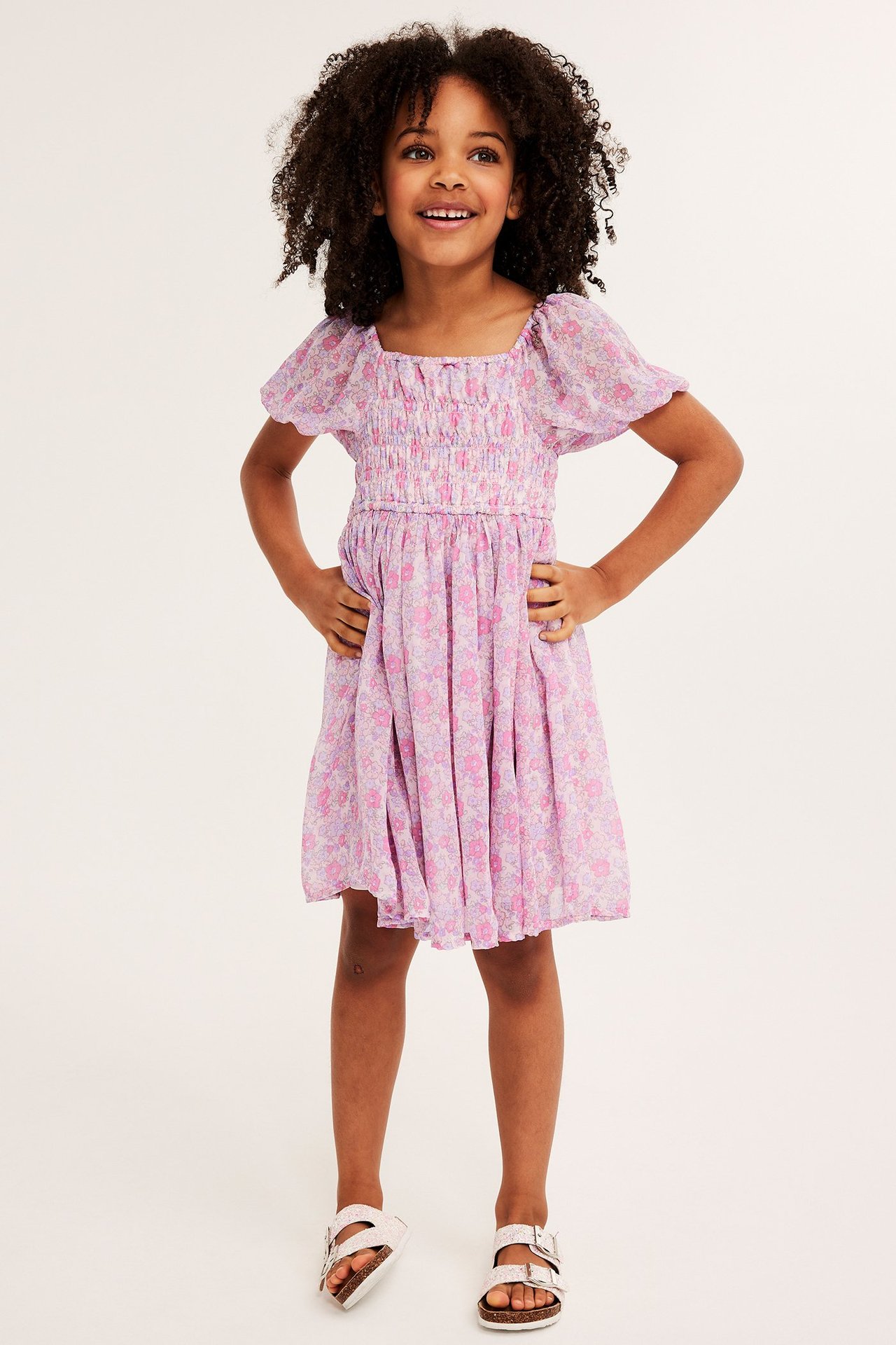 Widok z przodu uśmiechniętego dziecka w liliowej kwiecistej sukience szyfonowej ze smockiem i białych sandałach.