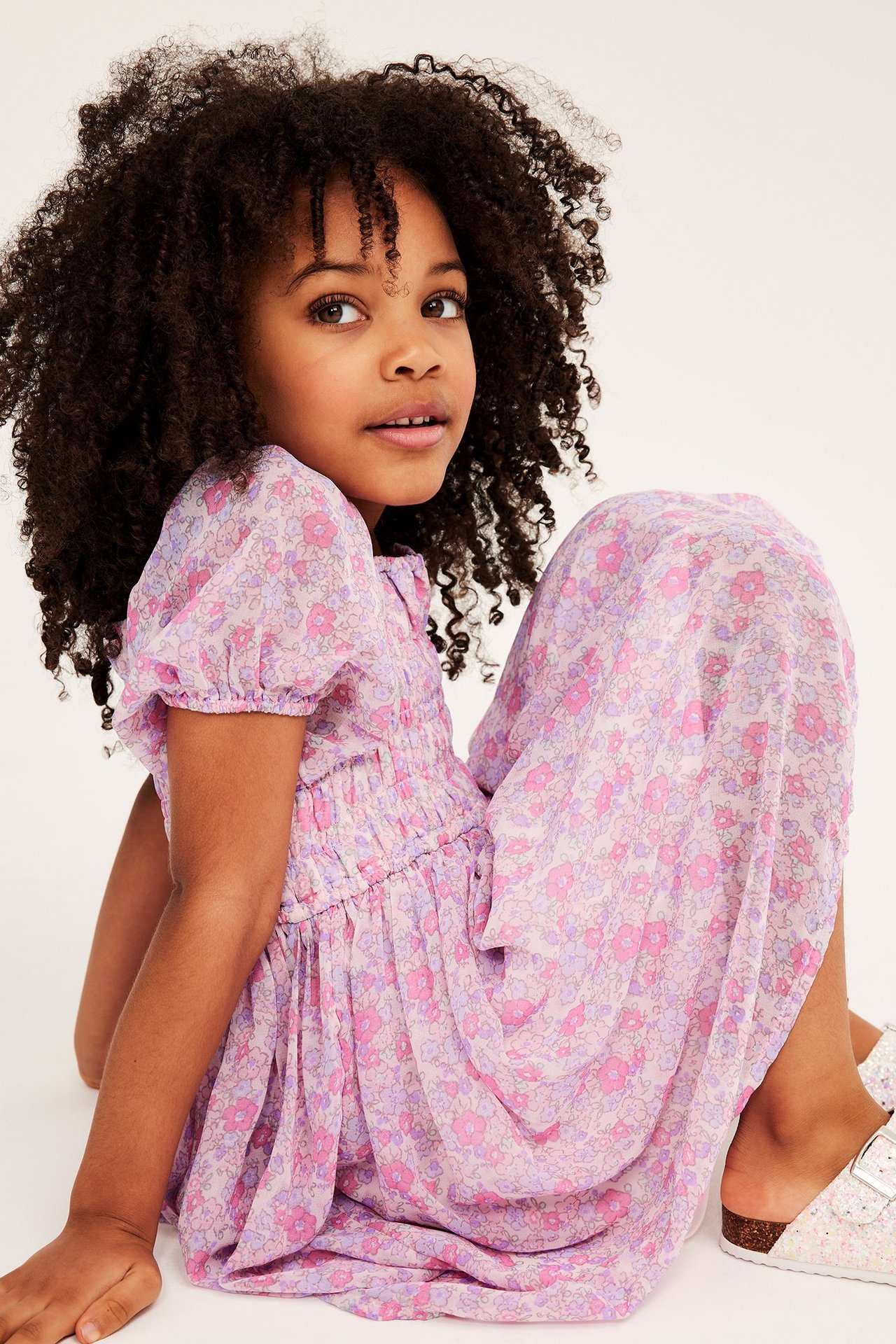 Widok z przodu: Dziecko w różowej kwiecistej sukience szyfonowej ze smockiem, bufiastymi rękawami i brokatowymi sandałami.