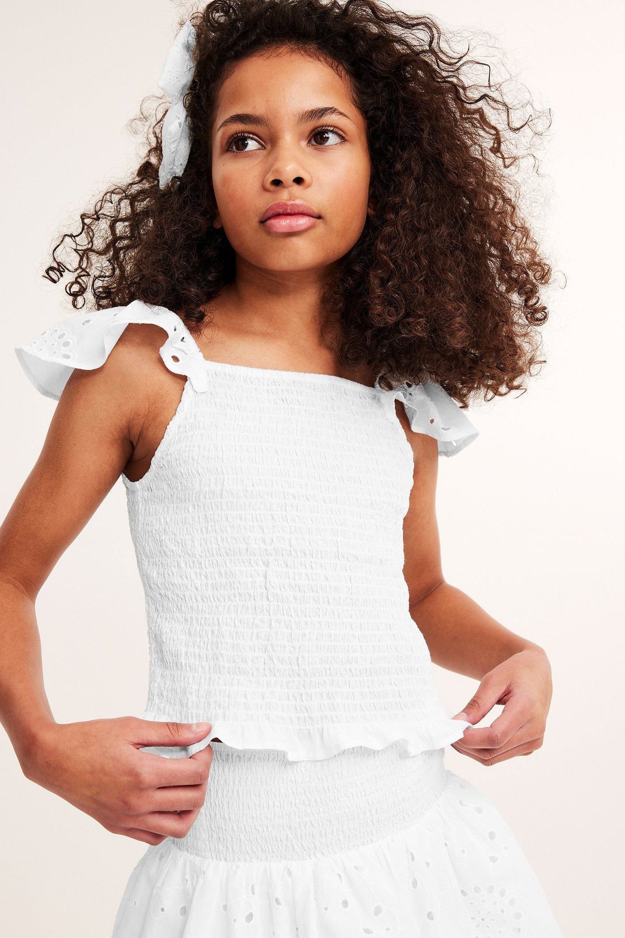 Frontbilde av et barn i en hvit smock-topp i bomull med broderie anglaise-volanger, matchende skjørt og hårsløyfe.