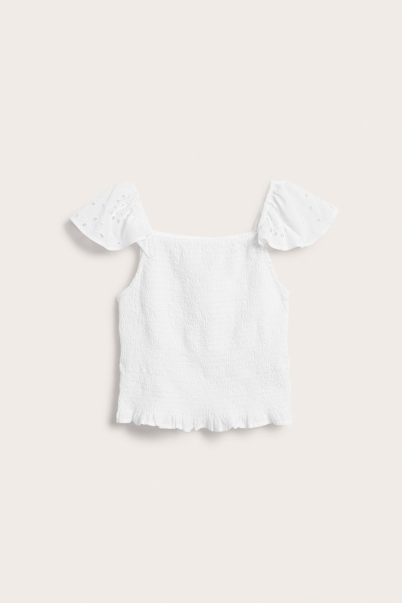 Frontvisning av en hvit smocket bomullstopp for barn med volangermer og kant i broderie anglaise.