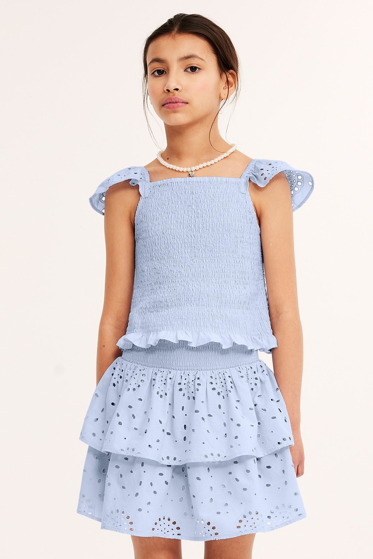Framifrån: Barn i ljusblå smockad topp med broderie anglaise-ärmar, matchande kjol och pärlhalsband.