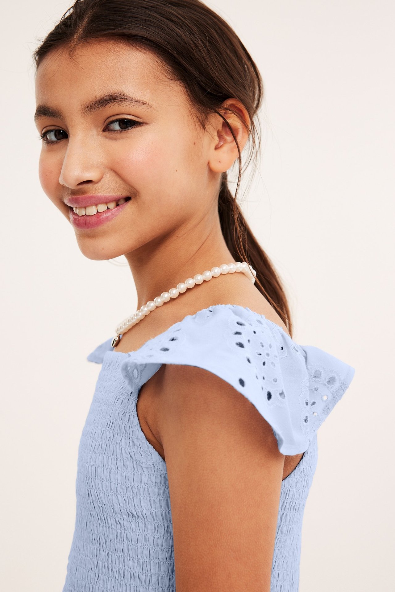 Zbliżenie, widok z boku dziecka w jasnoniebieskim marszczonym topie z falbankami broderie anglaise i naszyjnikiem z pereł.