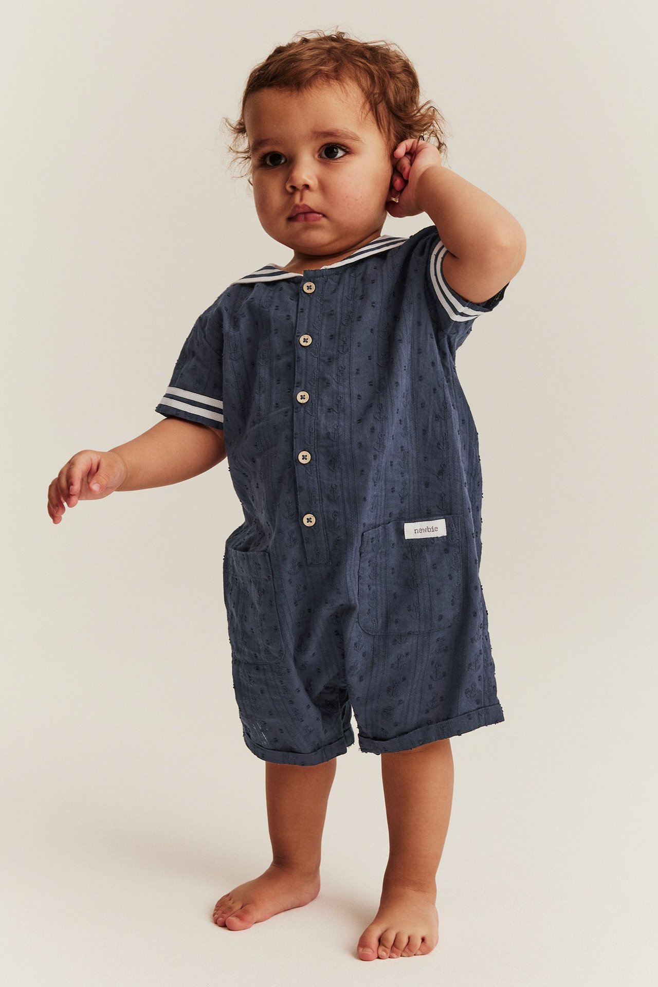 Edestä näkymä vauvasta tummansinisessä kuviollisessa puuvillahaalarissa, jossa on merimieskaulus ja napit edessä.
