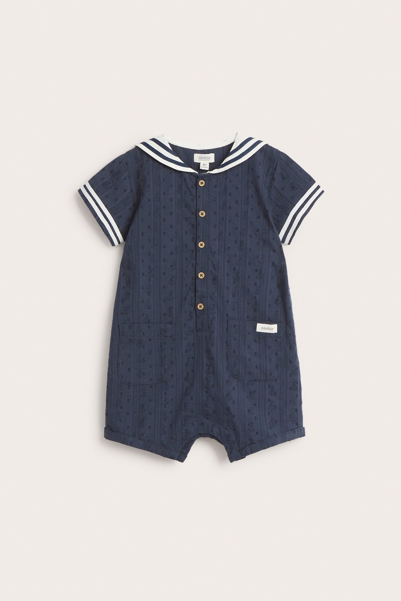Edestä: vauvan tummansininen kuviollinen puuvillahaalari valkoisella merimieskauluksella ja raidallisilla lyhyillä hihoilla.