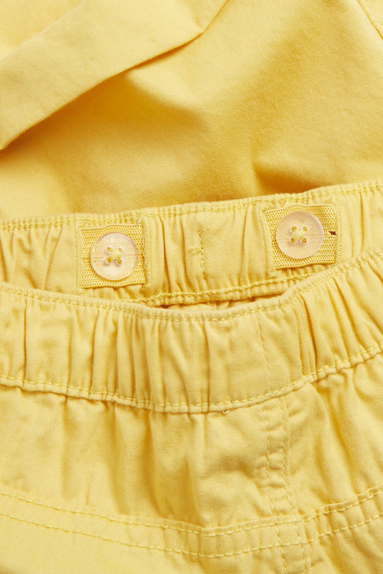 Zbliżenie żółtych bawełnianych szortów z elastyczną, regulowaną talią i dwoma widocznymi guzikami.
