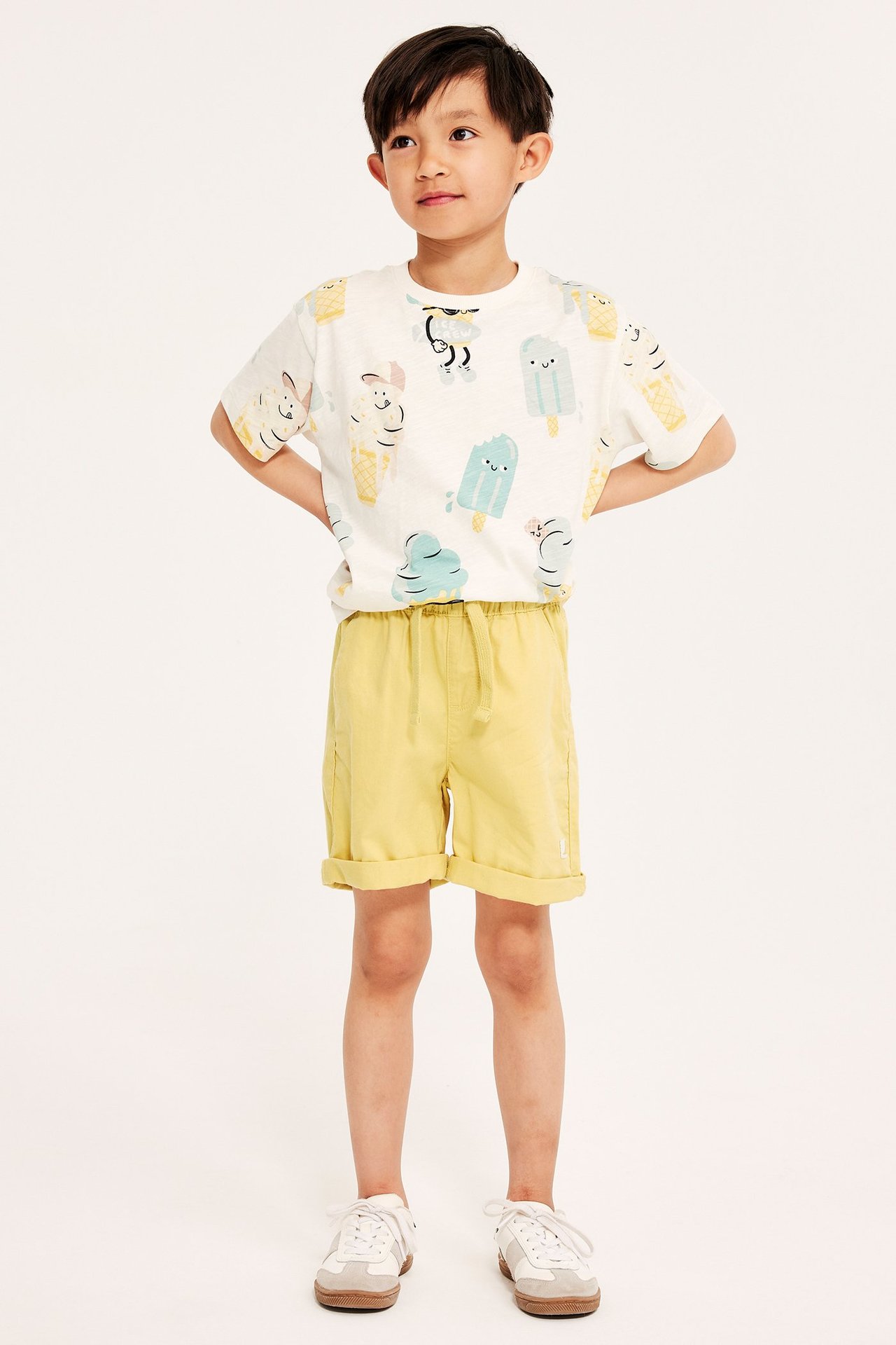 Widok z przodu: dziecko w żółtych bawełnianych szortach z gumką i białej koszulce z nadrukiem lodów.