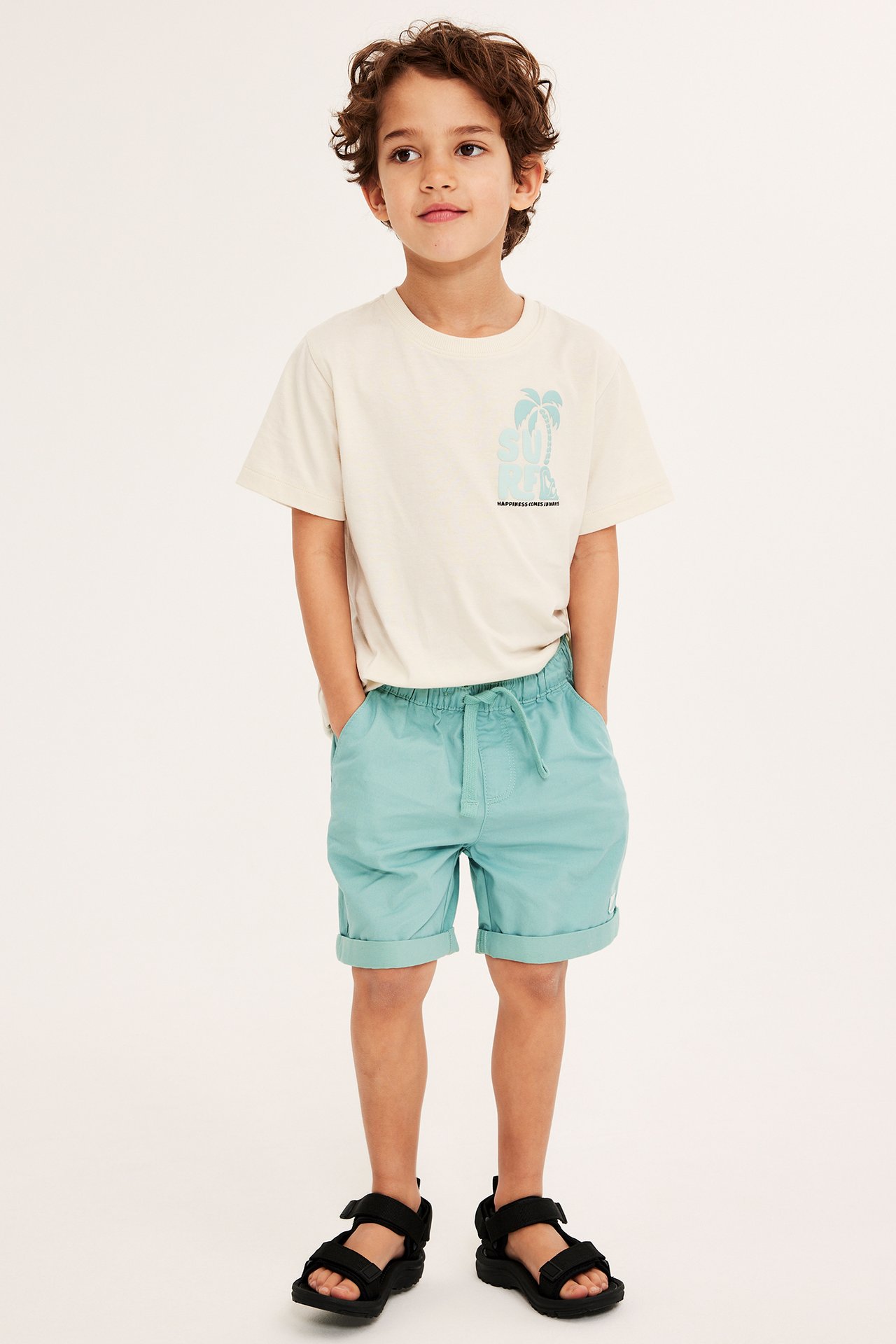 Frontvisning av et barn i lysegrønne bomullsshorts, en off-white grafisk t-skjorte og svarte sandaler.