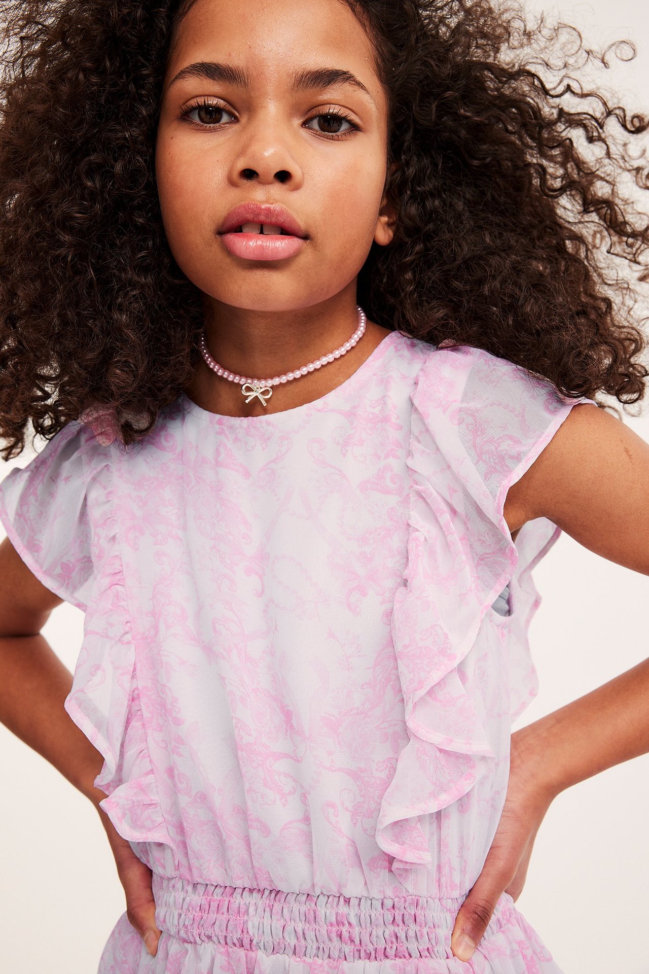 Nærbilde forfra av et barn i rosa mønstret chiffonkjole med volanger og rosa perlekjede med sløyfe.