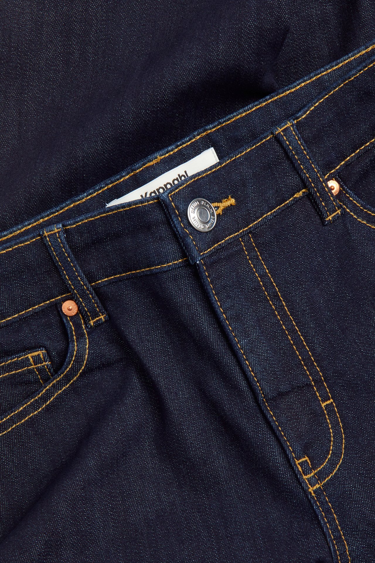 Lähikuva edestä tummansinisistä denim caprihousuista keltaisilla tikkauksilla, hopeanapilla ja etutaskuilla.