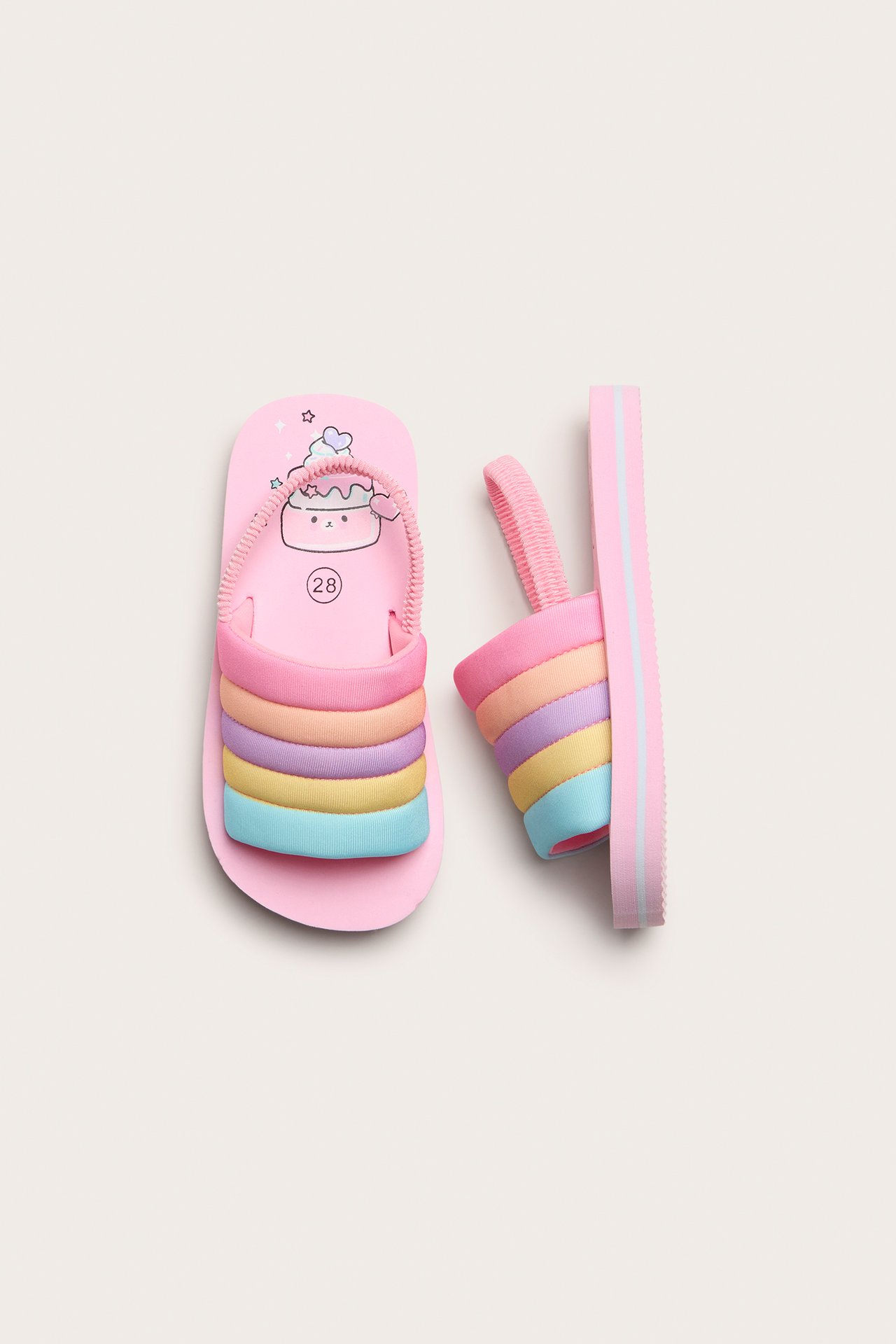 Uppifrån, rosa barnsandaler med regnbågsrandiga polyesterremmar och elastisk hälrem.