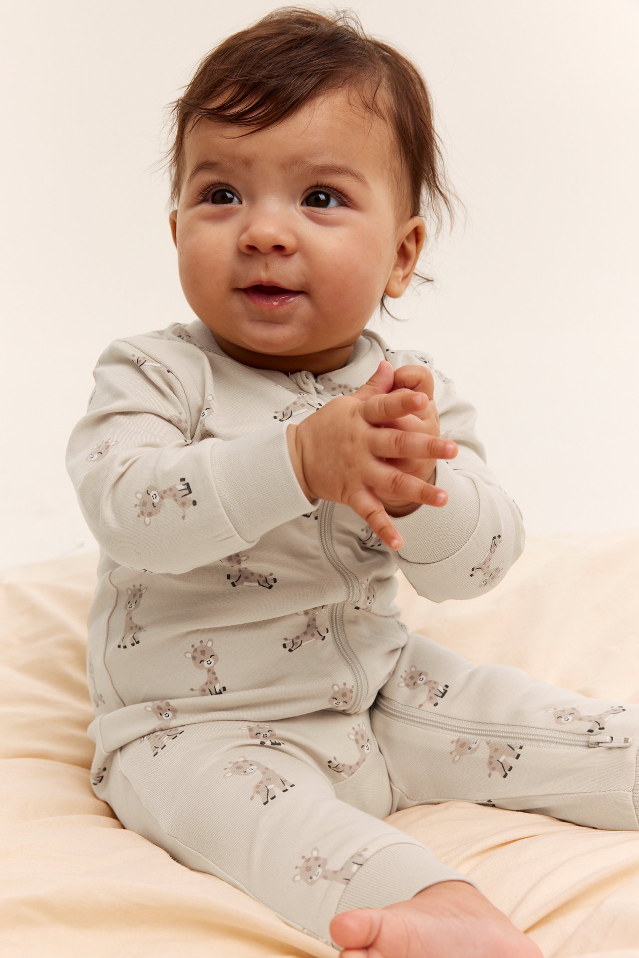 Framifrån: baby i offwhite bomullsromper med giraffmönster och tvåvägsdragkedja.
