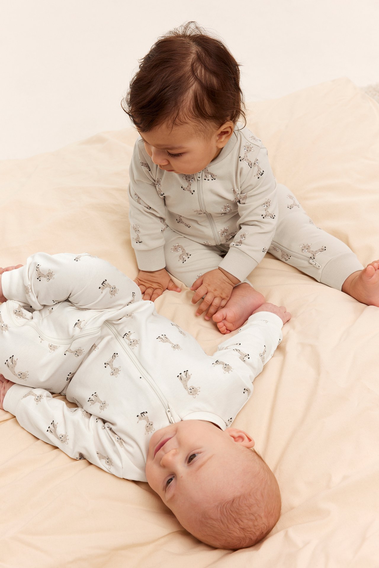 Framifrån, två bebisar i matchande offwhite djurmönstrade pyjamasar med tvåvägsdragkedja.