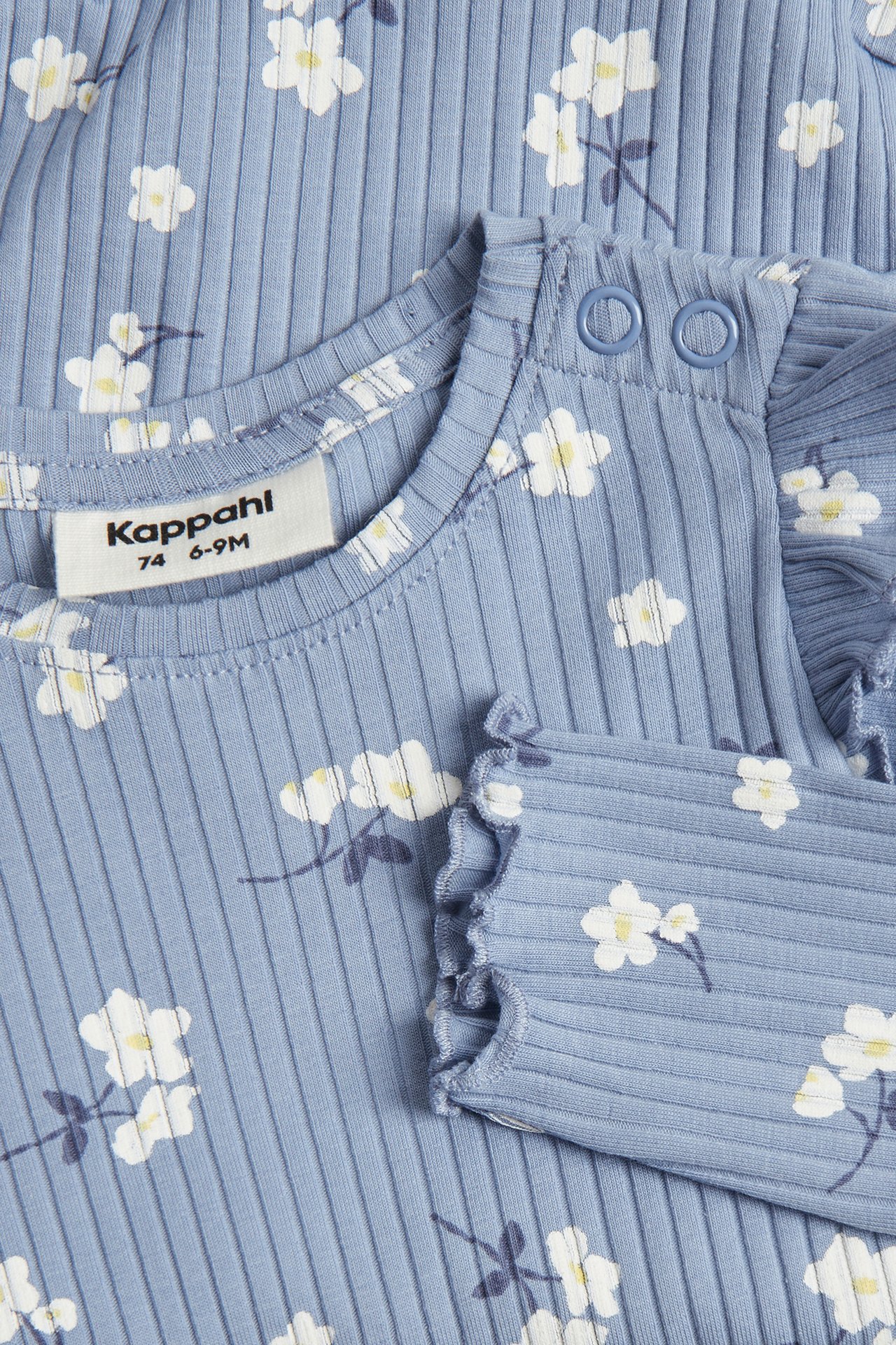 Närbild av en ljusblå ribbad babybody med vitt blommönster, axelvolanger och tryckknappar.
