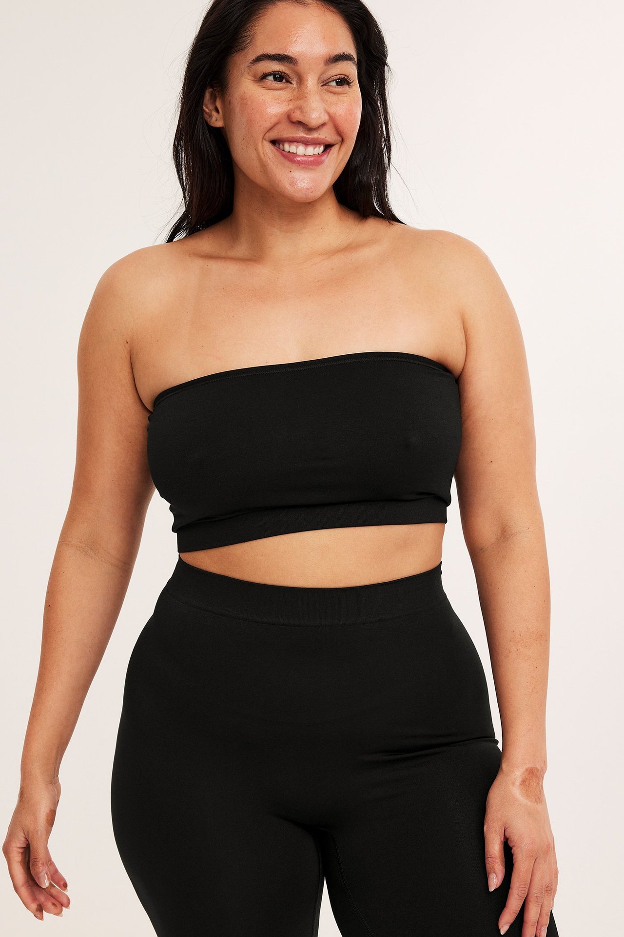 Framifrån, leende kvinna i svart sömlös bandeau-bh och svarta sömlösa högmidjade leggings.