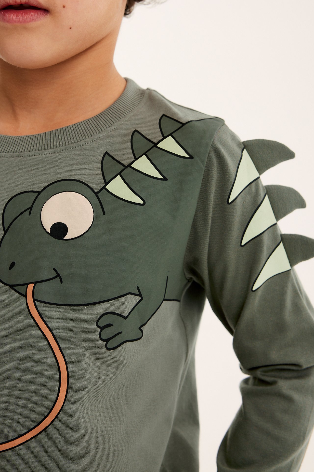 Närbild framifrån på ett barn i grön långärmad bomulls-t-shirt med ödletryck och applikationer på ärmen.