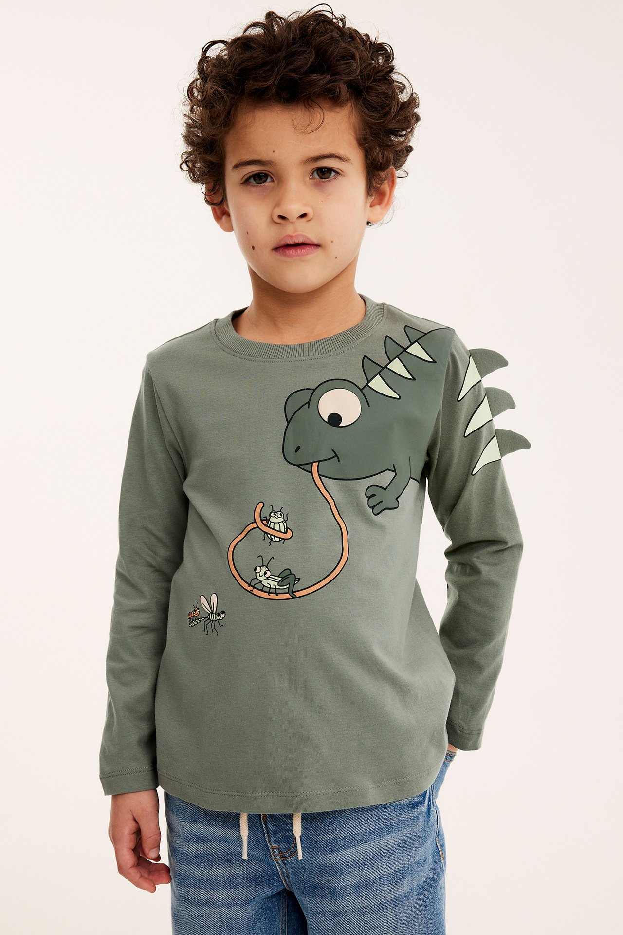 Framifrån, ett barn i grön långärmad bomulls-t-shirt med ödla- och insektsmönster, och blå jeans.