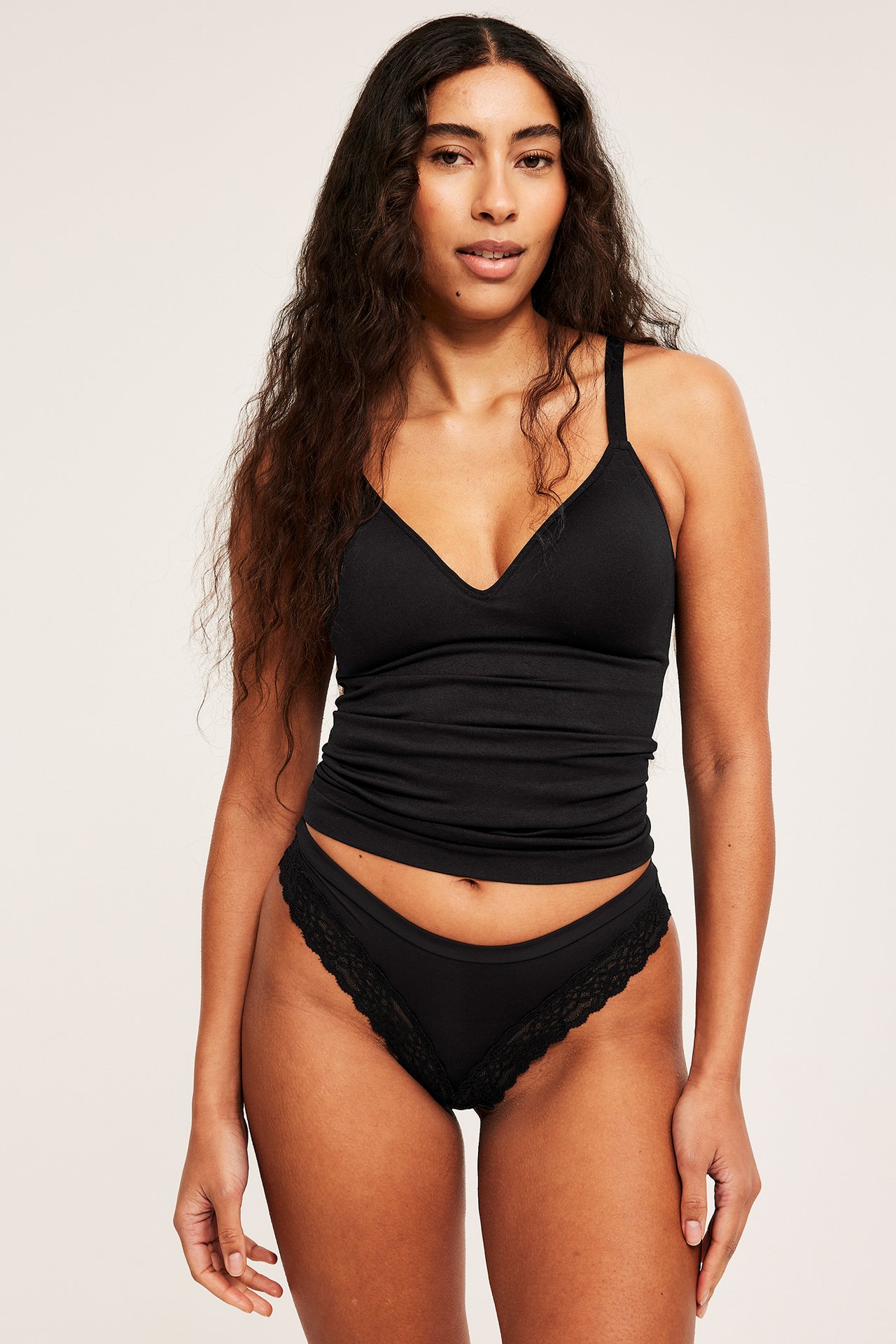 Edestäpäin, nainen yllään musta saumaton toppi sisäänrakennetuilla rintaliiveillä ja mustat pitsireunaiset alushousut.