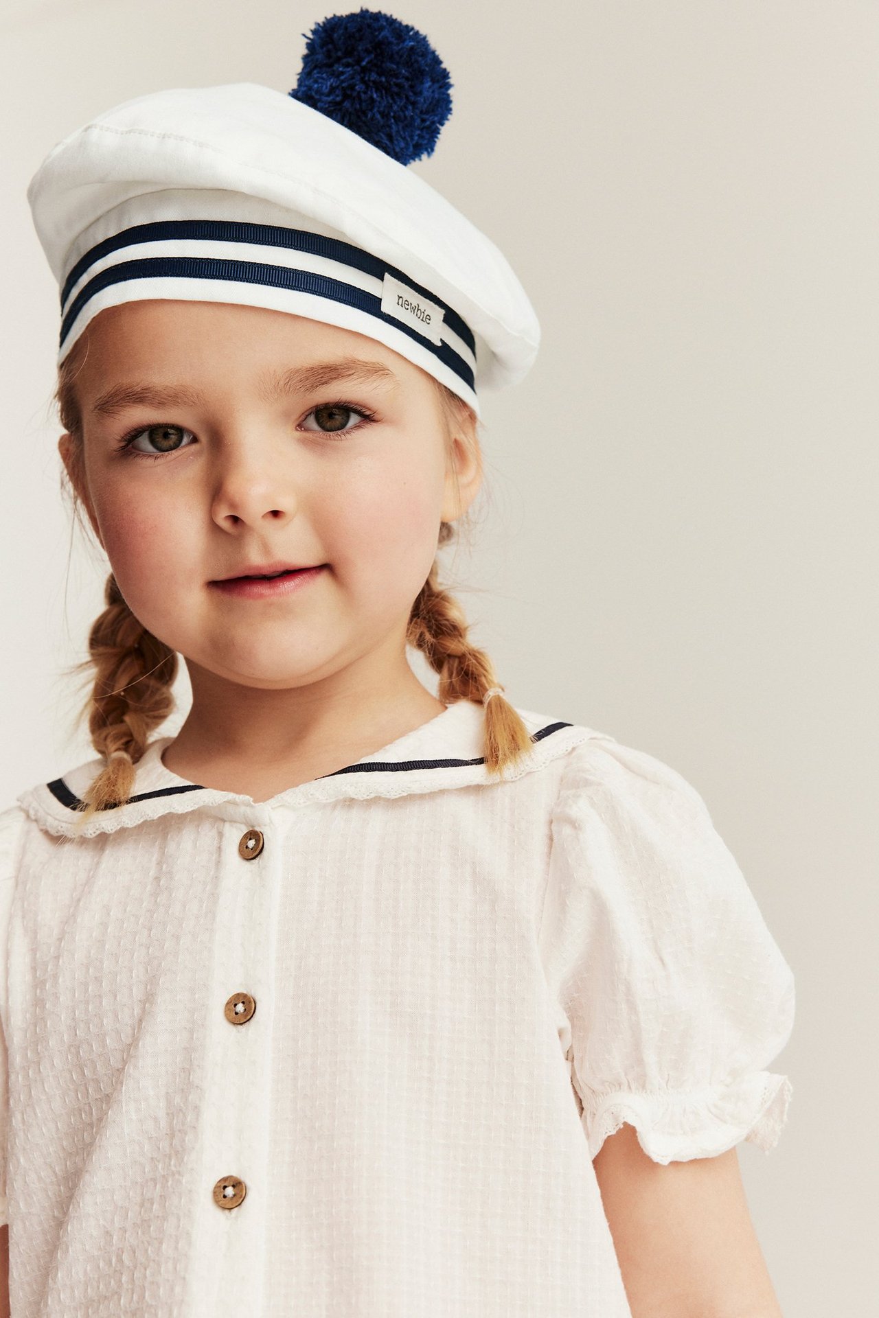Widok z przodu dziecka w kremowym bawełnianym berecie z niebieskimi paskami i pomponem, na teksturowanej bluzce.