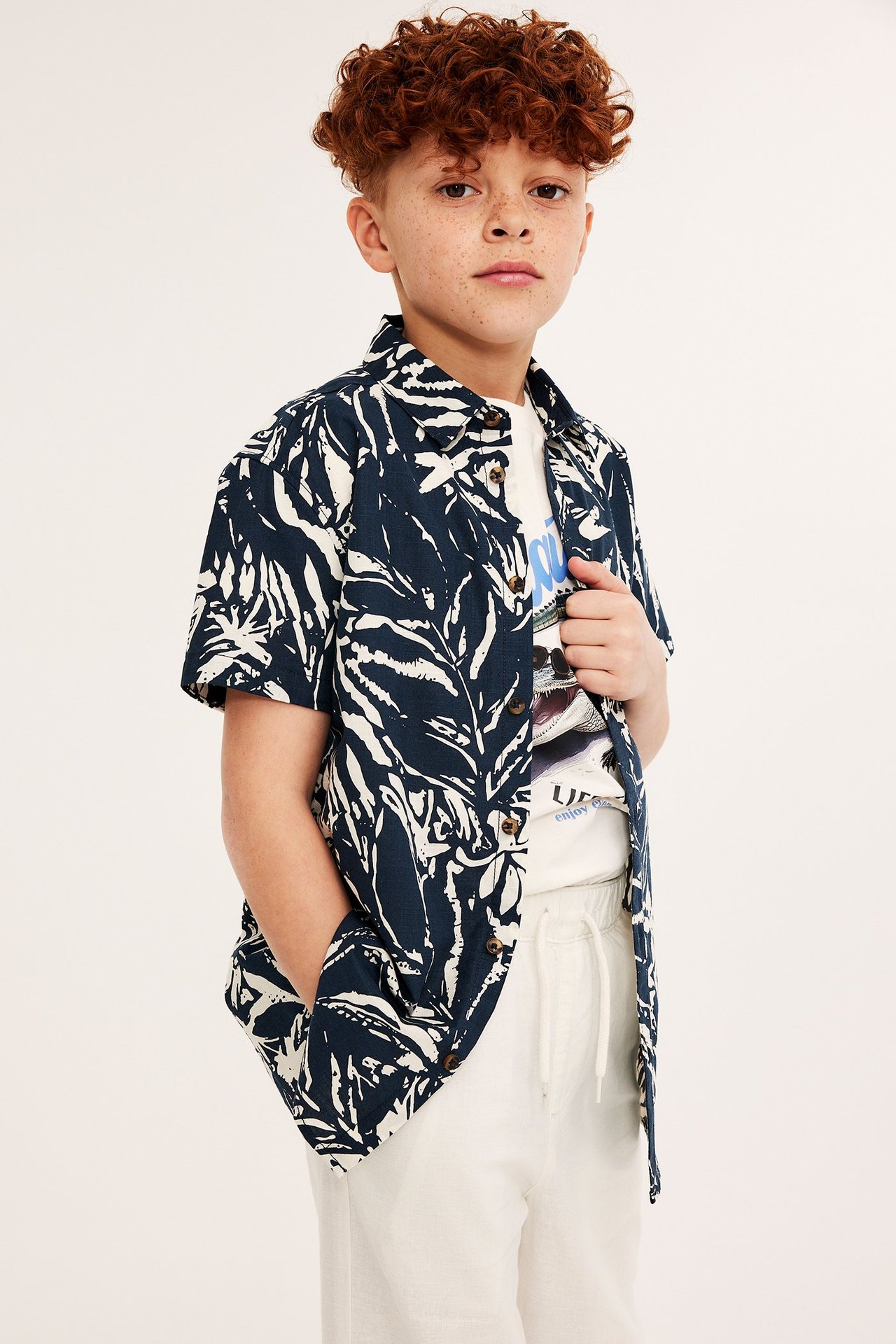 Widok z przodu: dziecko w granatowej wzorzystej koszuli z krótkim rękawem, białym T-shircie i jasnobeżowych szortach.