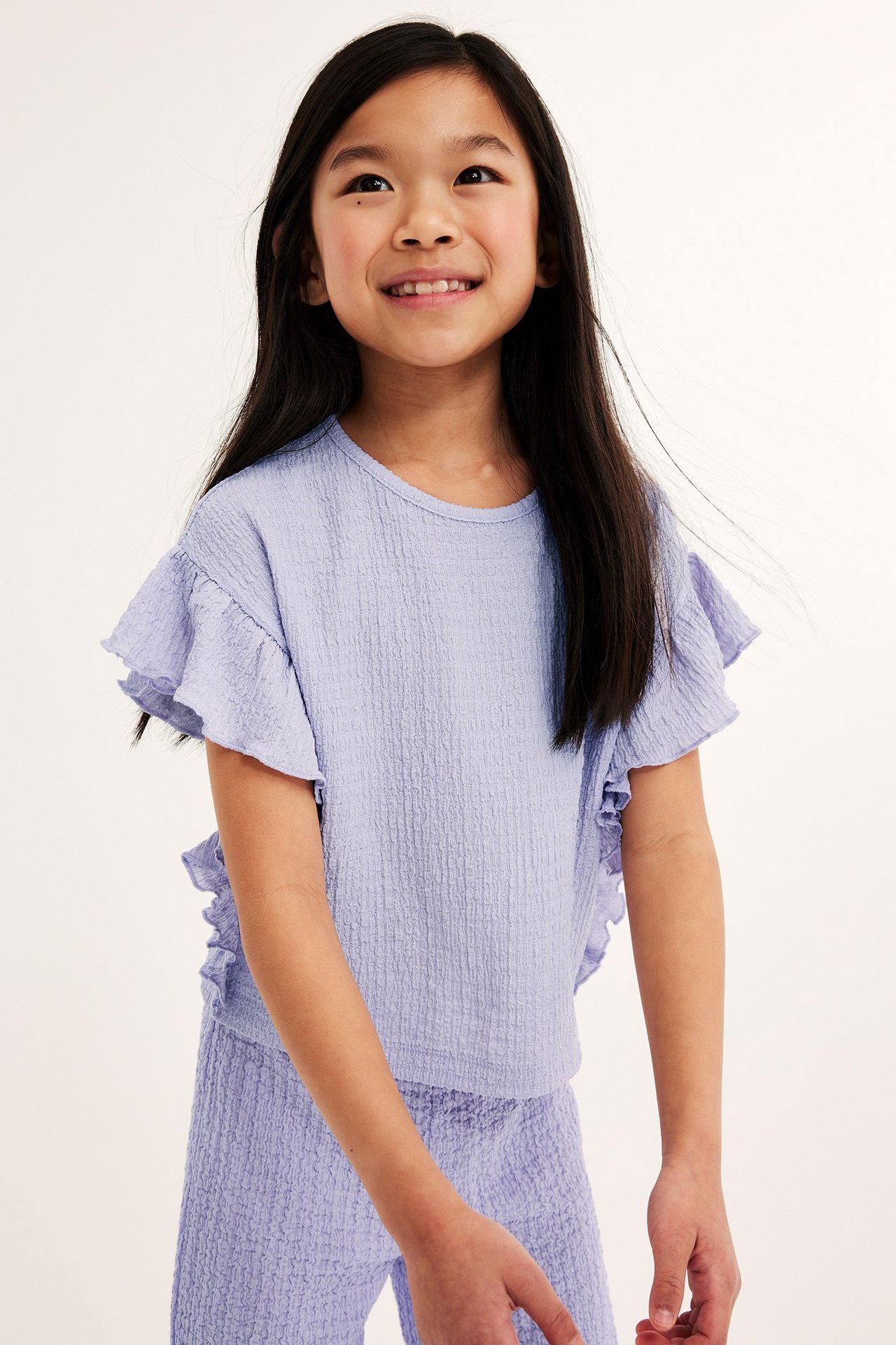 Widok z przodu dziecka w liliowym teksturowanym topie z falbankami i pasujących liliowych spodniach.