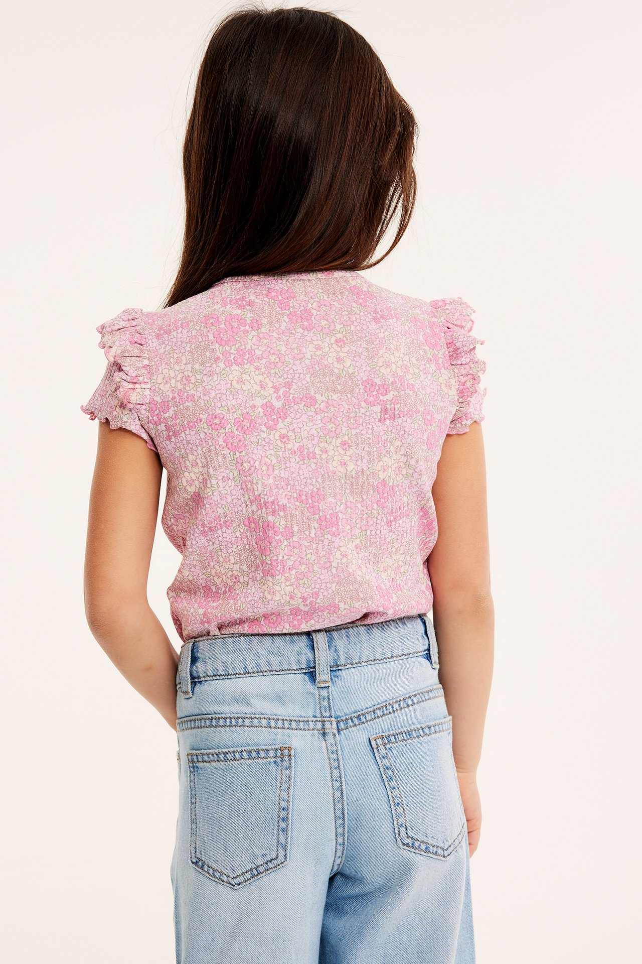 Bakfra, et barn i lys rosa blomstret ribbet topp med volangermer og lyseblå jeans.