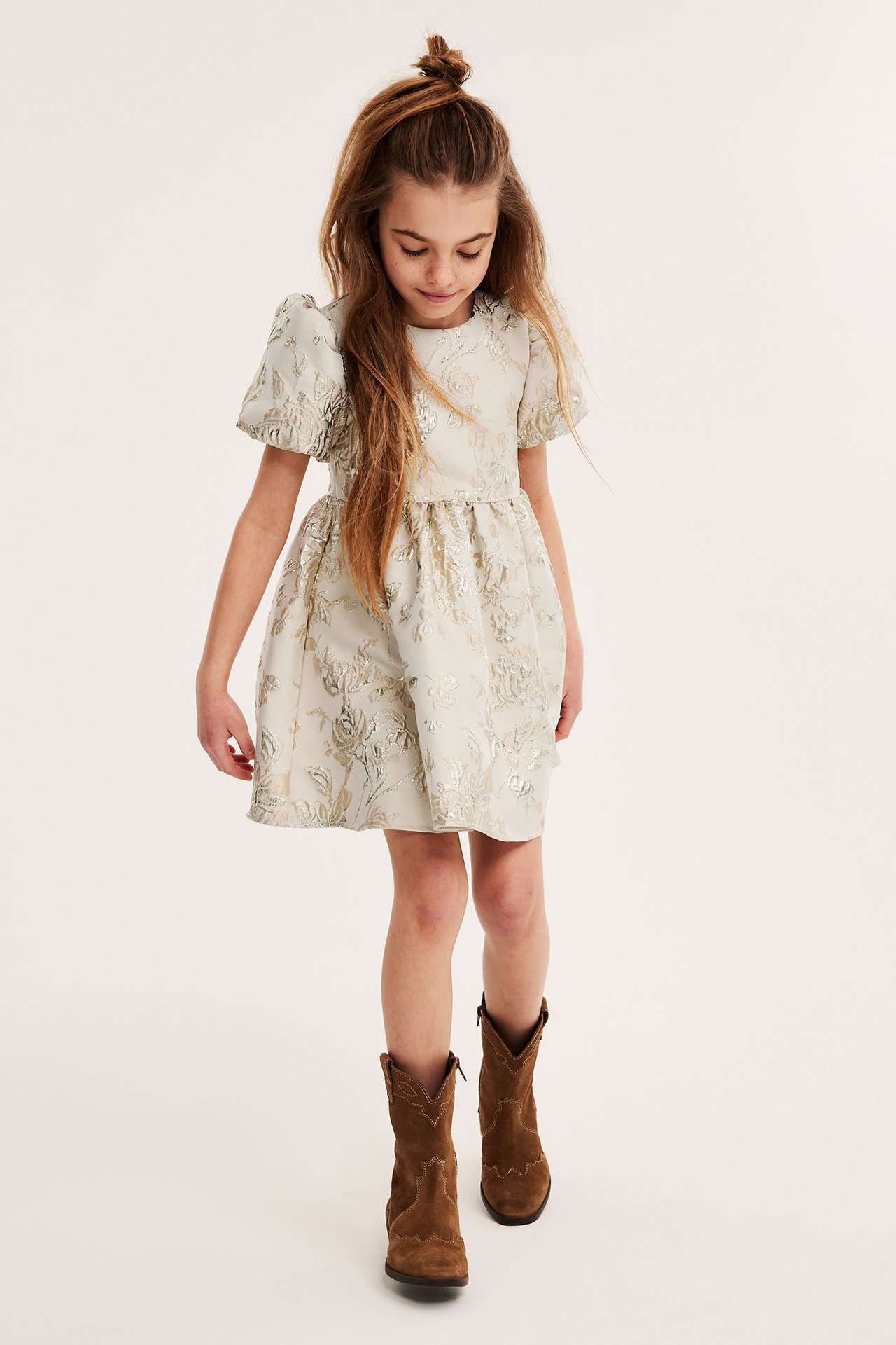 Widok z przodu dziecka w jasnozłotej brokatowej sukience z bufiastymi rękawami i brązowych zamszowych kowbojkach.