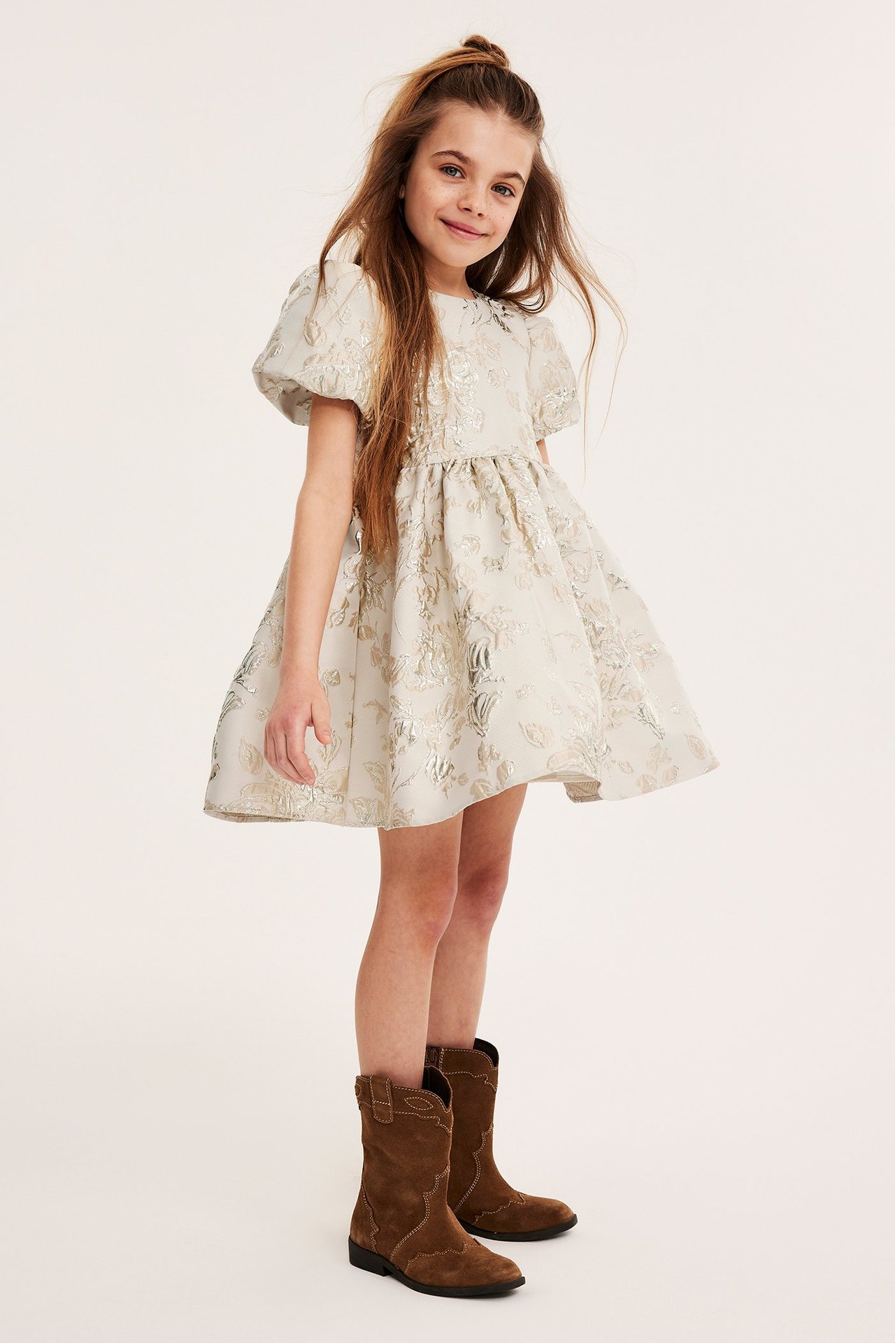 Widok z przodu dziecka w złotej żakardowej sukience brokatowej z bufiastymi rękawami i brązowych zamszowych butach.