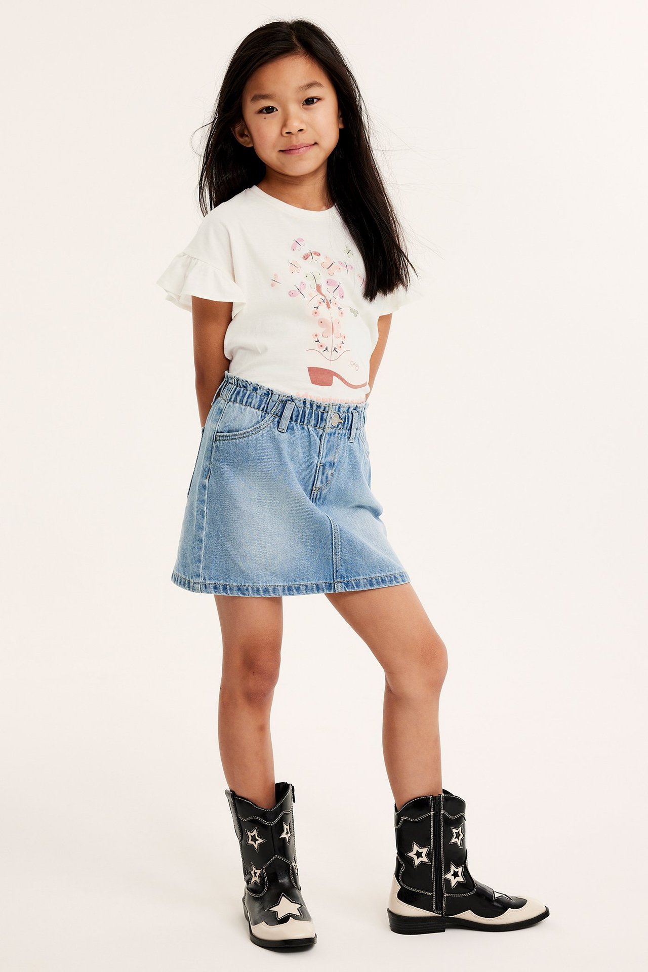 Widok z przodu: Dziecko w białym topie z falbankami, nadruk buta, jasnoniebieska spódnica jeansowa, czarne kowbojki w gwiazdki.