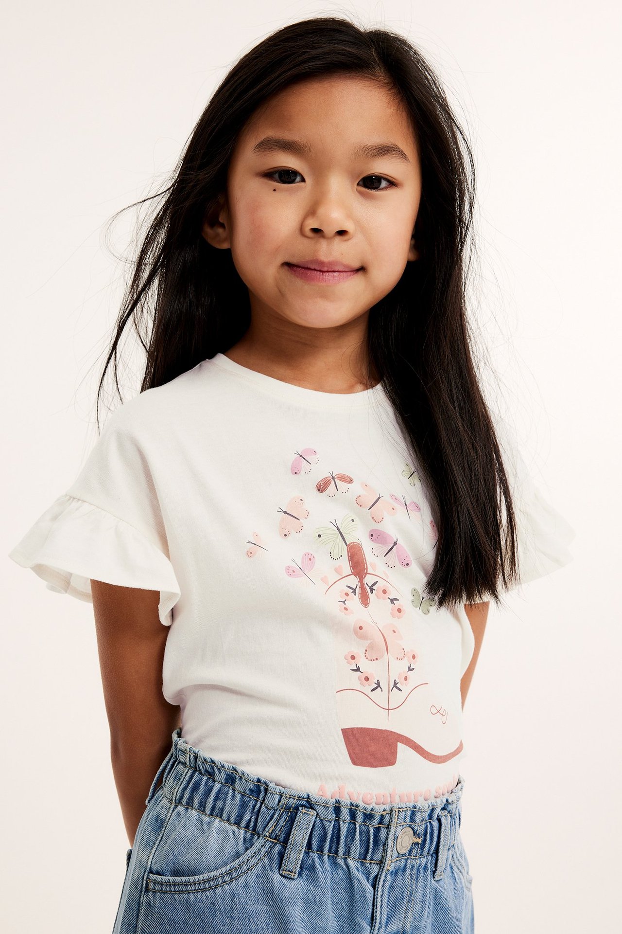 Widok z przodu dziecka w ecru bawełnianym topie z falbankami i nadrukiem motyli, w połączeniu z niebieskimi dżinsami.