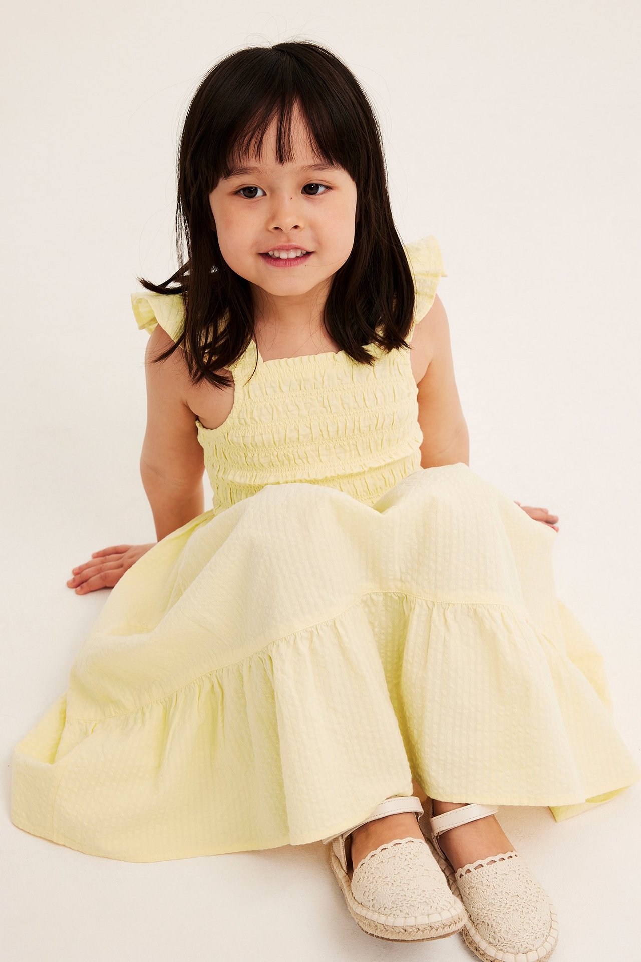 Widok z przodu: Dziecko w jasnożółtej sukience z seersuckeru ze smockowym gorsetem, falbankami i kremowymi espadrylami.