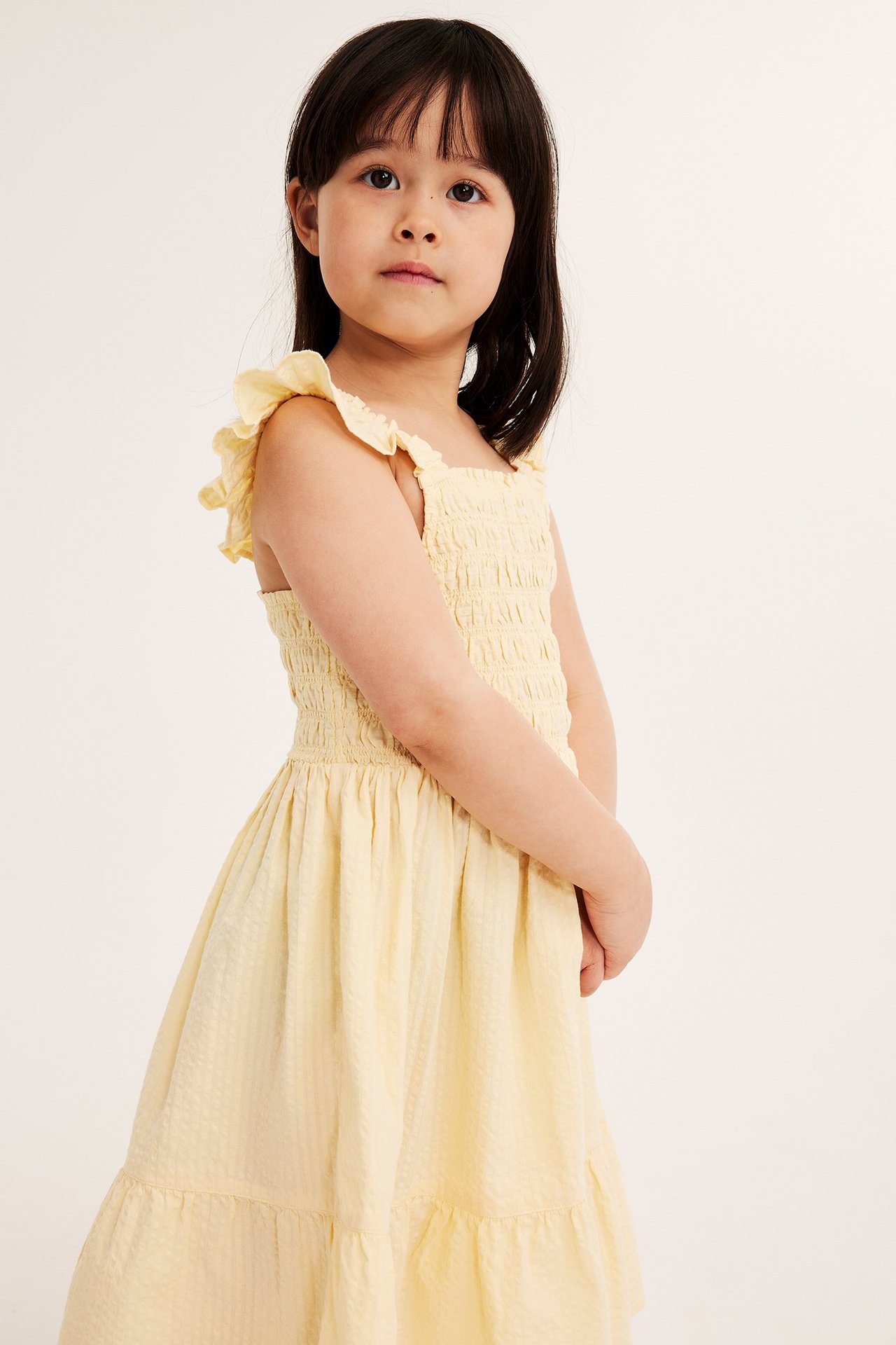 Widok z przodu dziecka w jasnobeżowej sukience z bawełny seersucker, ze smockowanym gorsetem i falbankami.