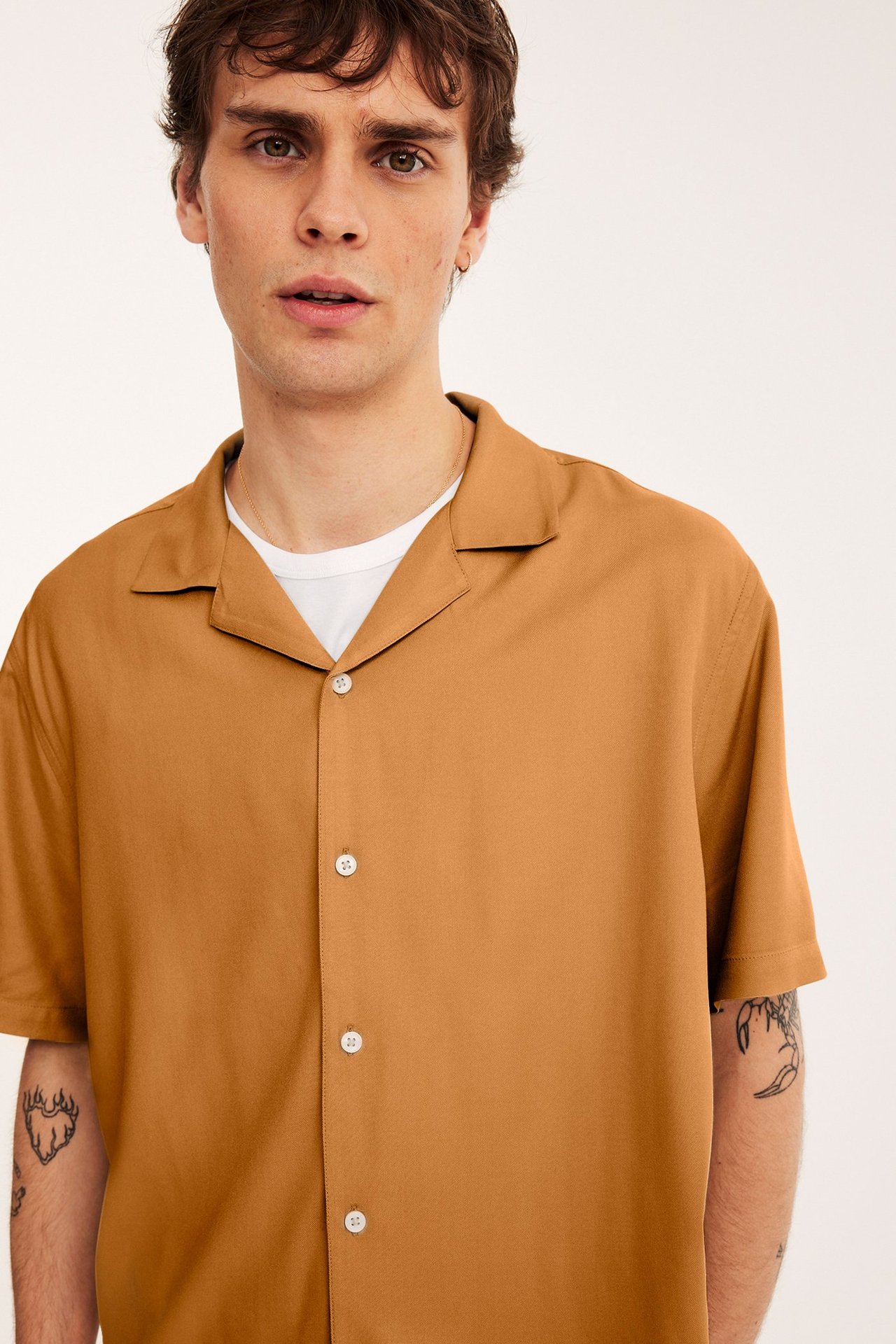 Edestäpäin, mies yllään ruskea lyhythihainen viskoosipaita valkoisilla napeilla valkoisen t-paidan päällä.