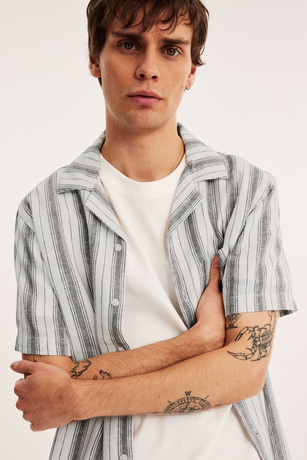 Edestä kuvattu mies yllään vaaleanharmaa/siniraitainen lyhythihainen pellavasekoitepaita, avoin valkoisen t-paidan päällä.