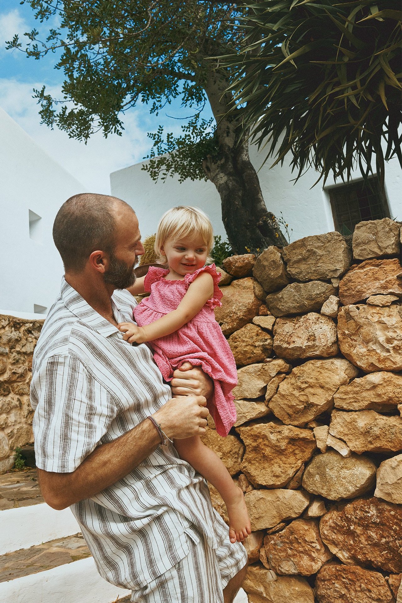 Edestä: Mies raidallisessa pellavasekoite-lyhythihaisessa paidassa ja shortseissa, pitää vauvatyttöä vaaleanpunaisessa mekossa.