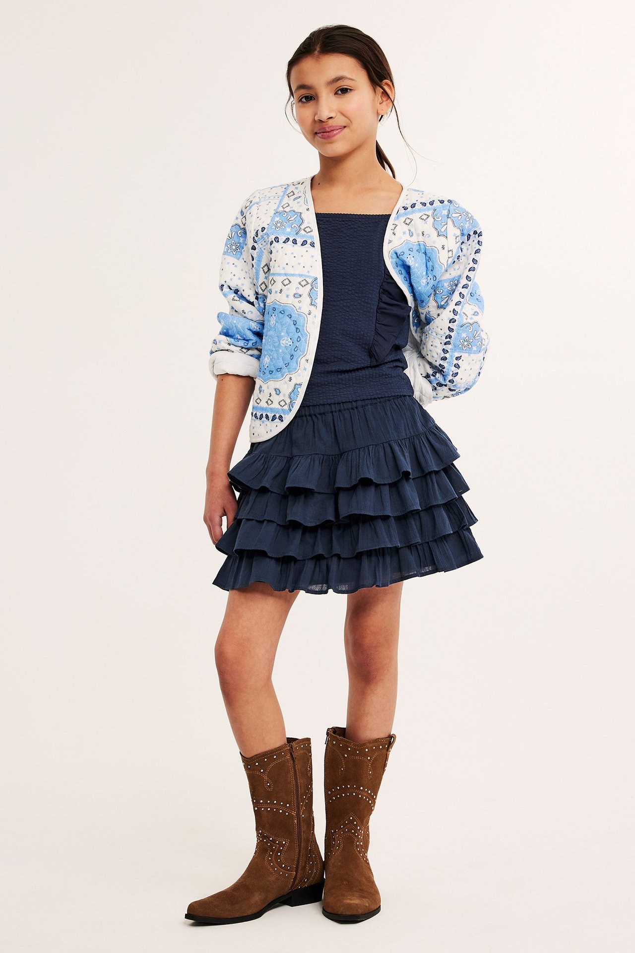 Forfra: et barn i mørkeblått bomulls-volangskjørt, marineblå topp, mønstret jakke og brune støvler.