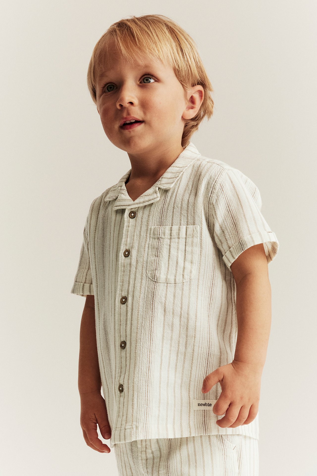Edestä: lapsi luonnonvalkoisessa raidallisessa lyhythihaisessa puuvillapaidassa ruskeilla napeilla ja shortseissa.