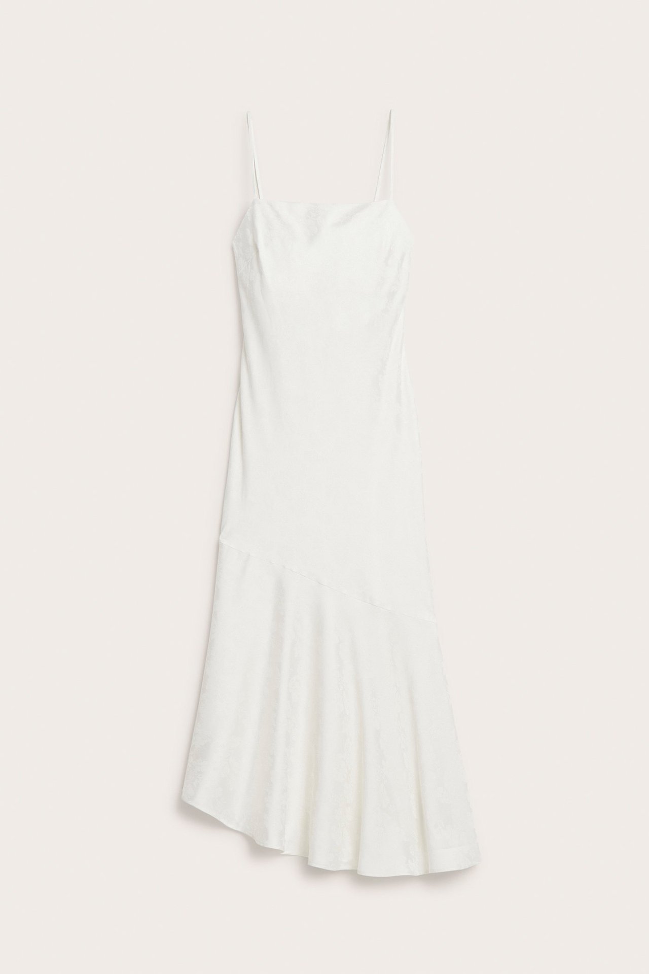 Frontvisning av en offwhite jacquard asymmetrisk slipkjole med spaghettistropper og utsvingt kant.