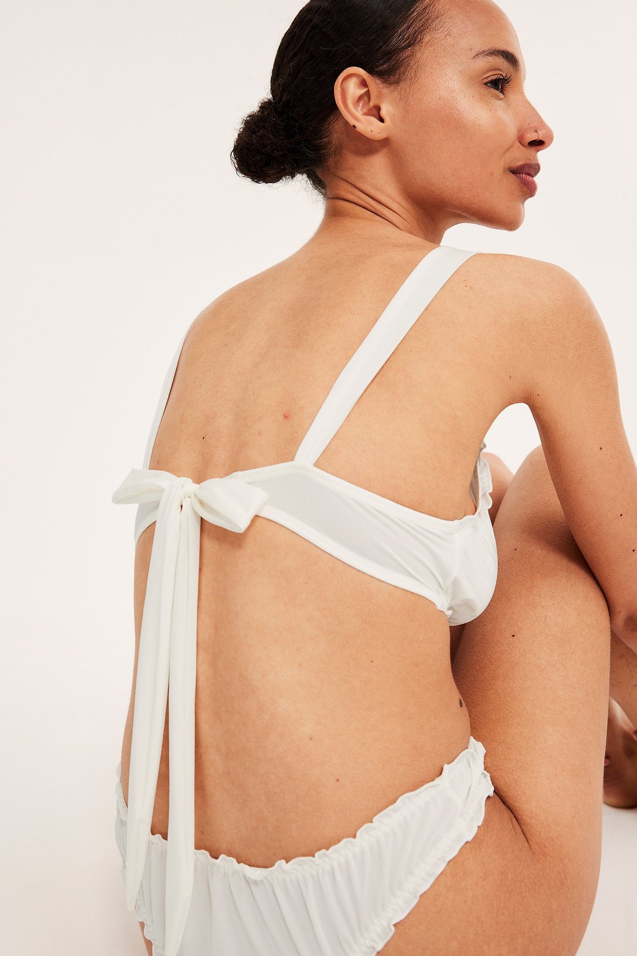 Bakifrån: en kvinna i en offwhite skimrande BH med dekorativ knytning bak och matchande volangtrosor.