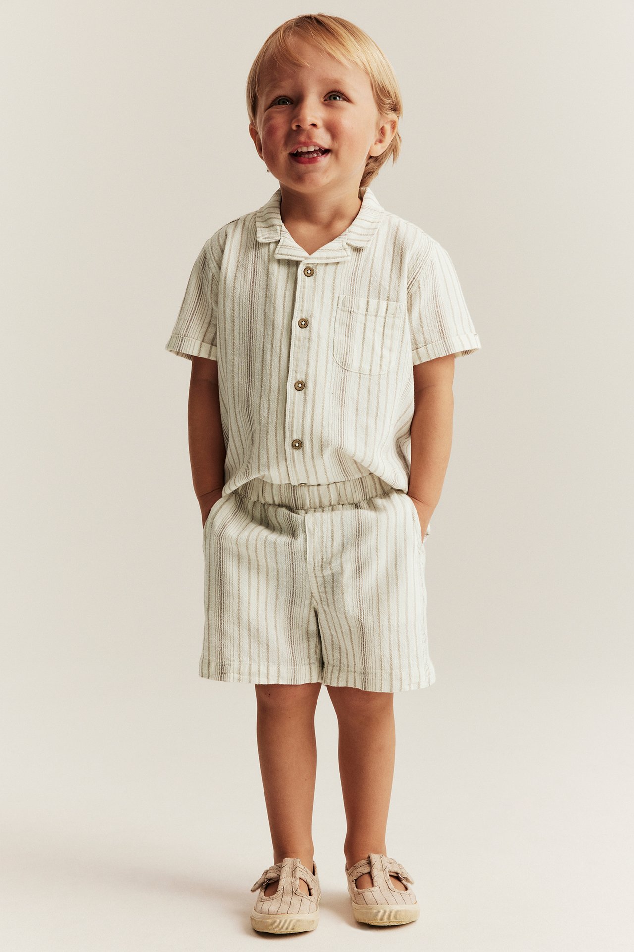 Frontbilde av et smilende barn i offwhite stripete vevde bomullsshorts, matchende skjorte og beige sko.