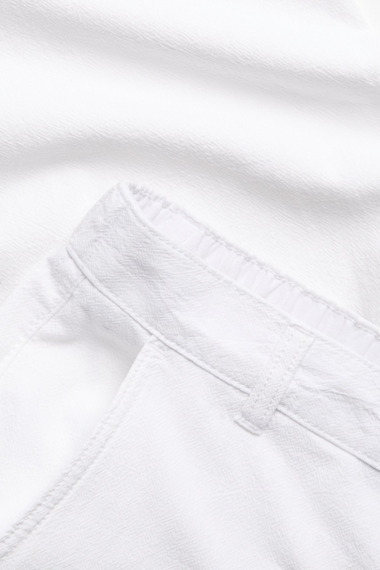 Nærbilde av hvite bomullsbukser i linstil med teksturert stoff, synlig linning og beltehemper.