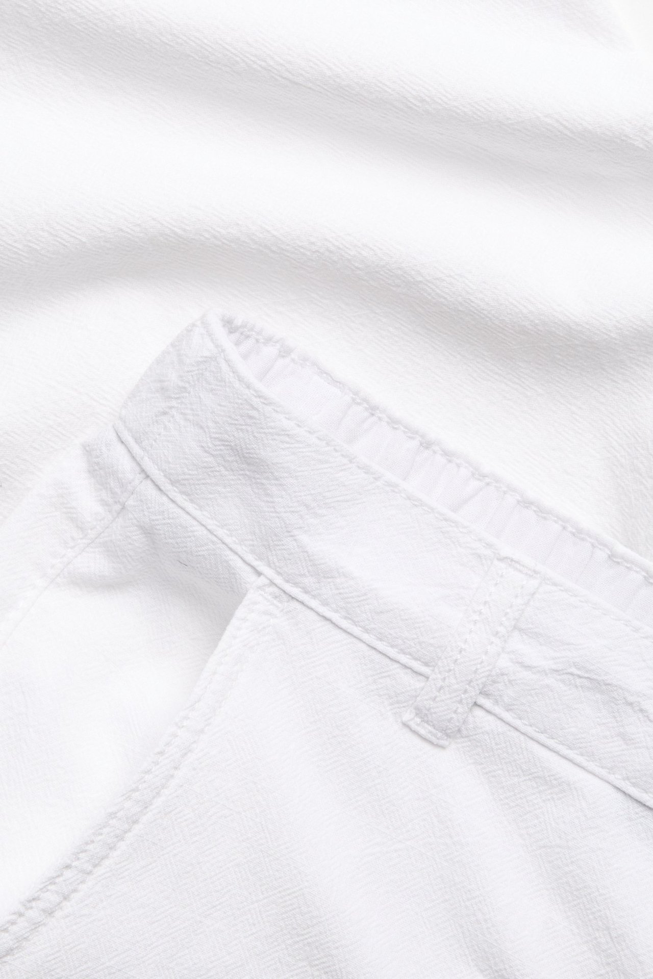 Zbliżenie na białe bawełniane spodnie o wyglądzie lnu, z teksturowaną tkaniną, widocznym pasem i szlufkami.