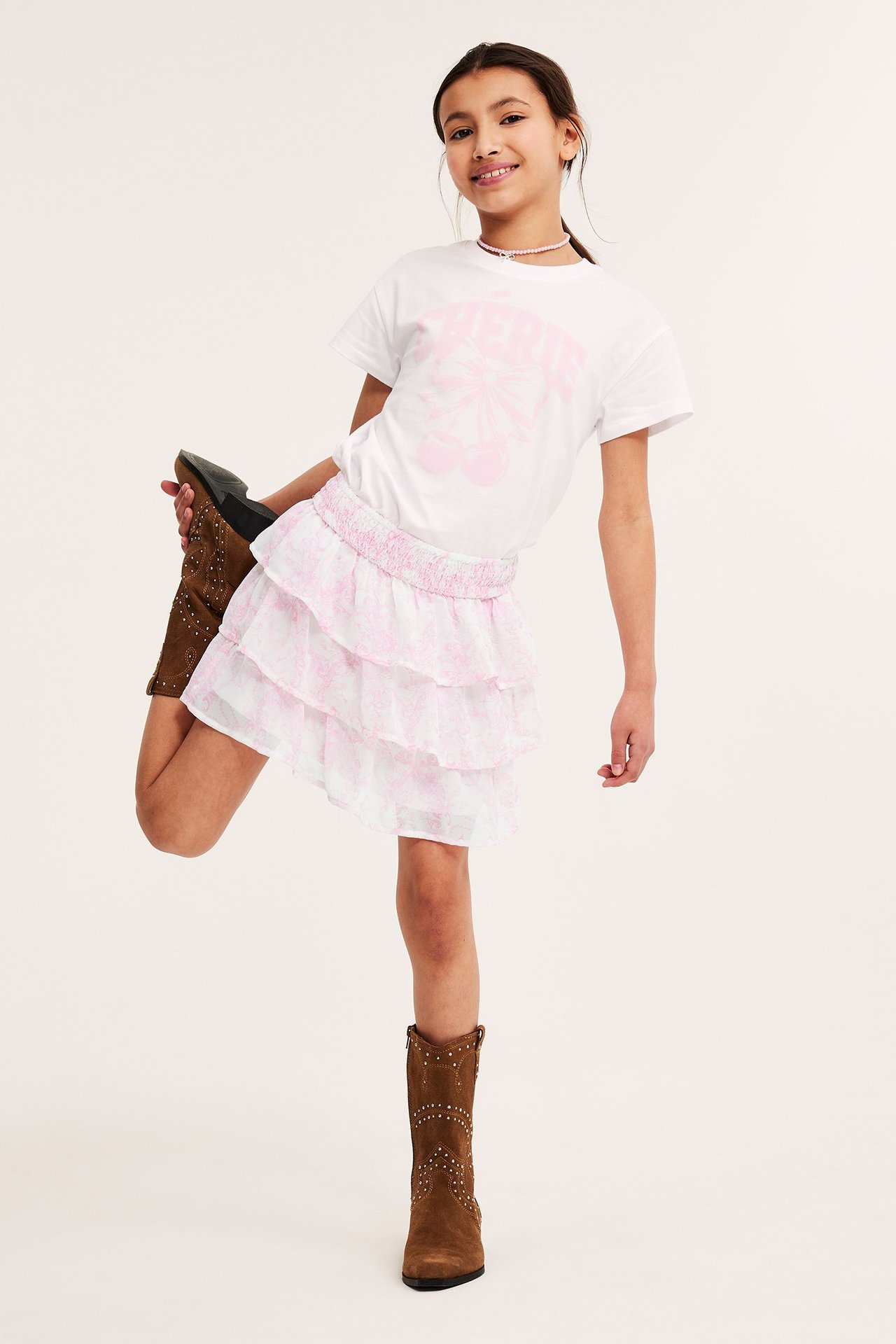 Framsida: Flickbarn i rosa mönstrad volangkjol i chiffong, vit t-shirt, bruna stövlar, rosa halsband.