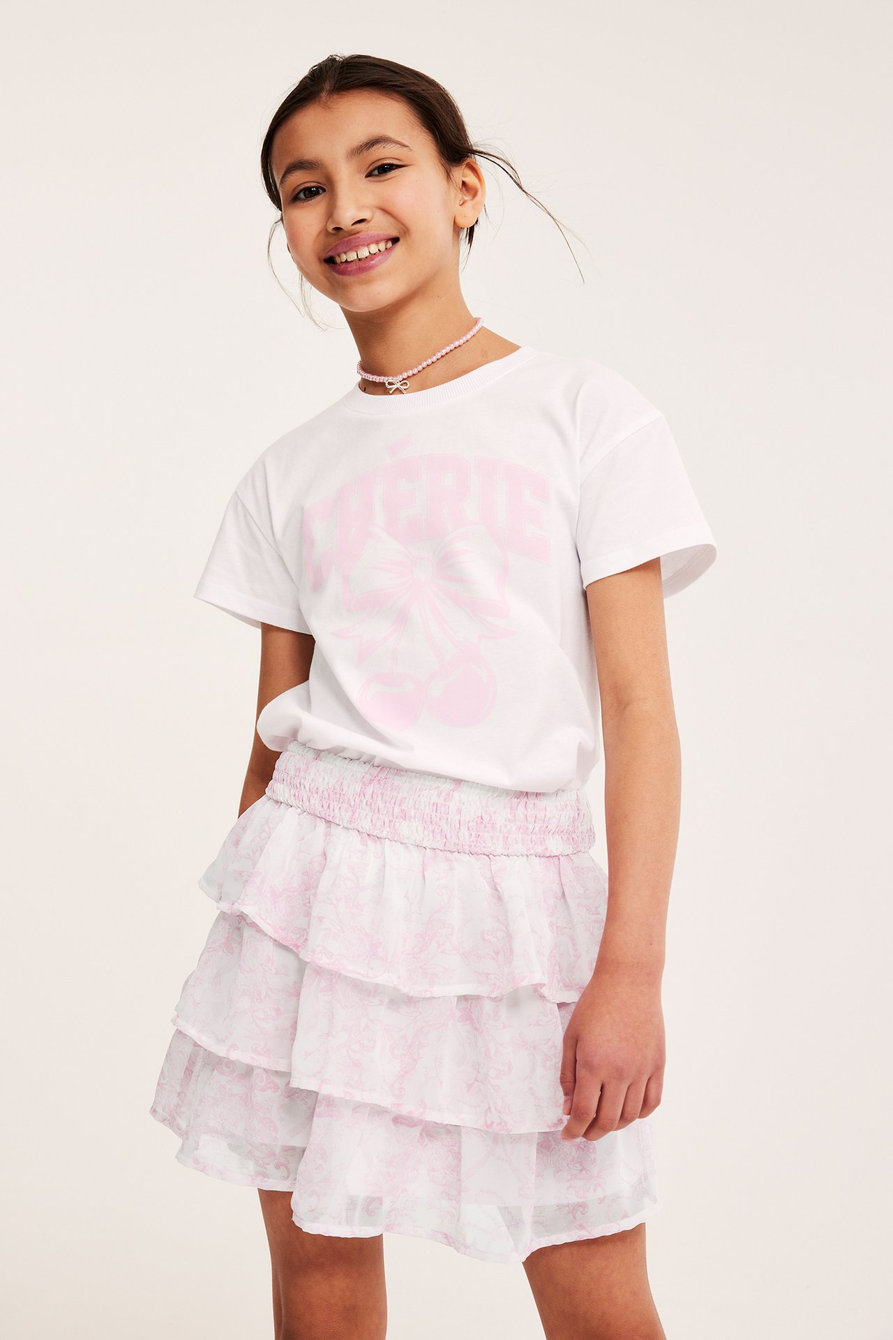 Framsida av ett barn i en rosa mönstrad volangkjol i chiffong, en vit t-shirt och ett rosa pärlhalsband.