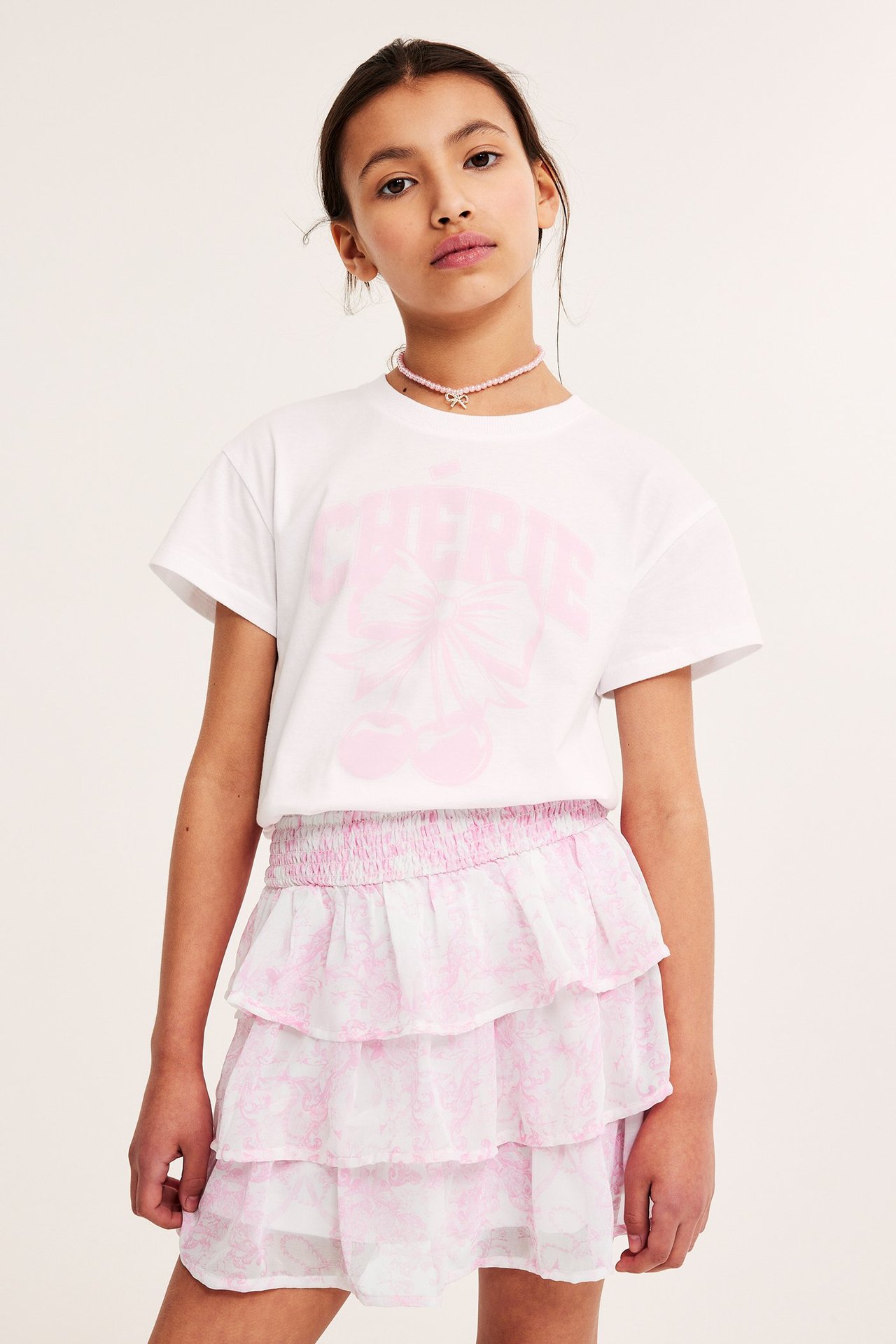 Frontvy av ett barn i en rosa mönstrad volangkjol i chiffong, vit t-shirt och rosa pärlhalsband.